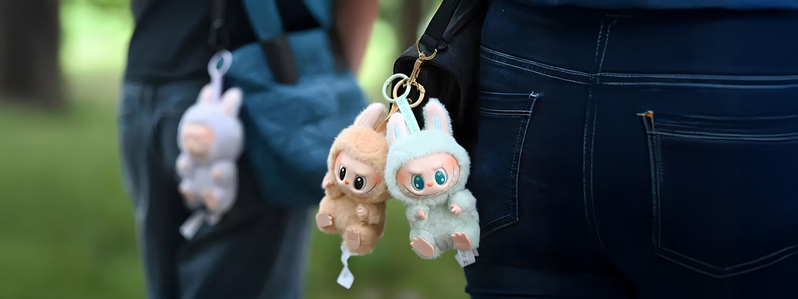 Two small Labubu plush keychains in tan and mint green with bunny ears clipped to a black bag strap against dark denim jeans in an outdoor setting.