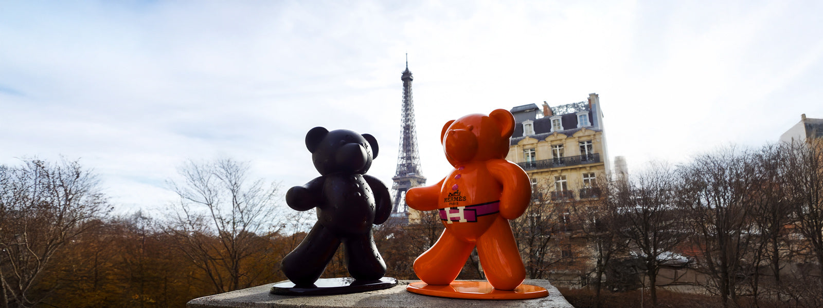 Two André Gacko bear sculptures in matte black and glossy orange with a pink Hermès-style belt facing each other on a ledge near the Eiffel Tower.