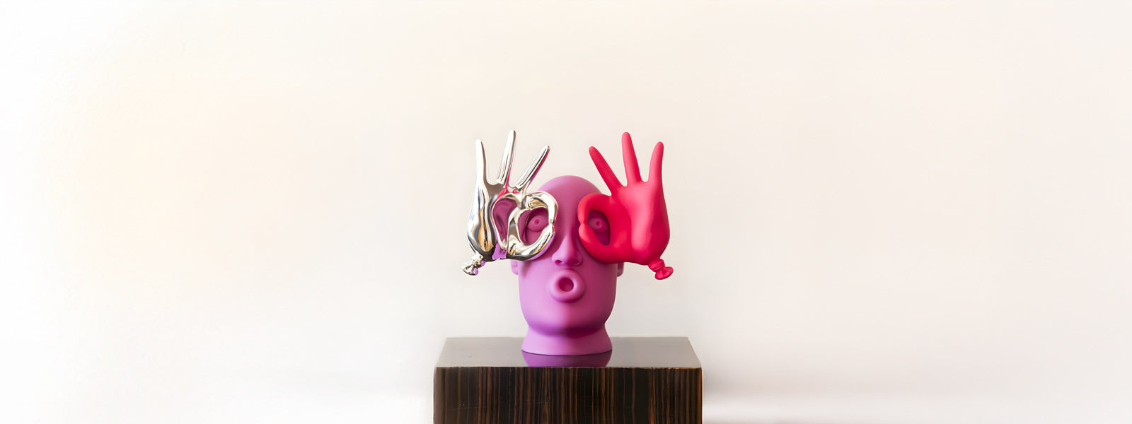 Purple pop art head sculpture with pursed lips, a chrome peace-sign hand over one eye and a red OK-sign hand over the other, on a dark wood pedestal.
