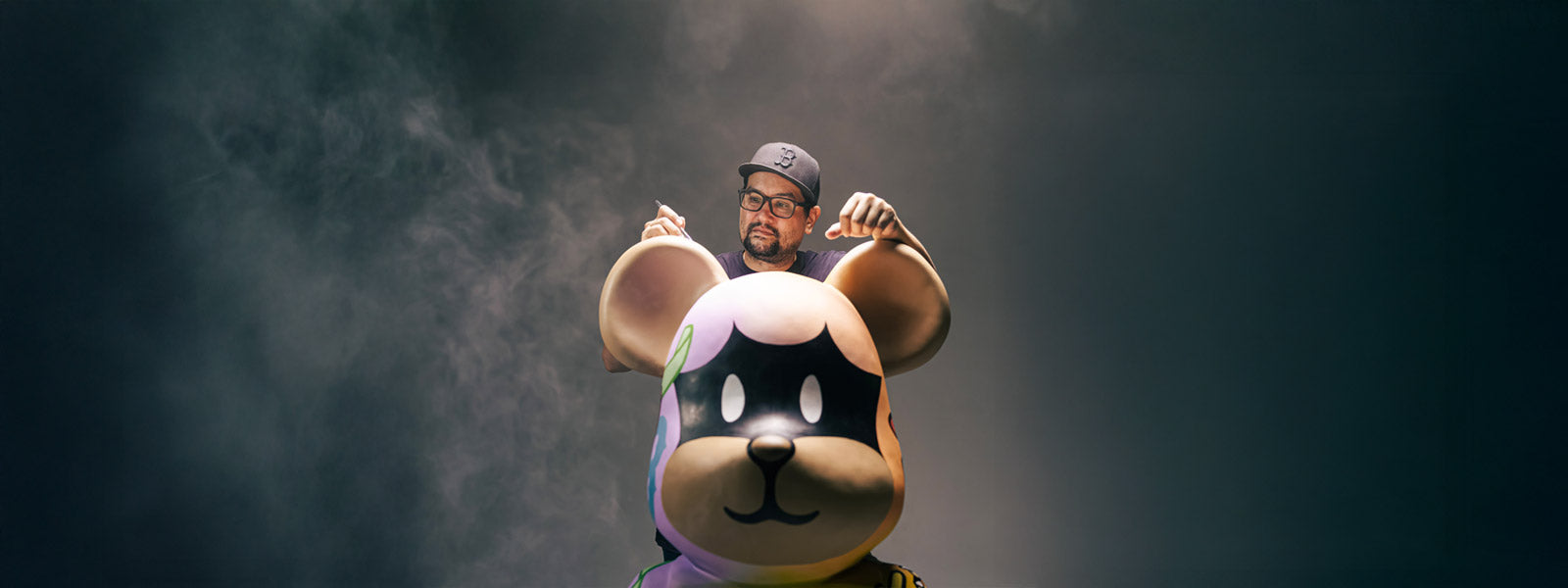 Artist BigDel posing behind a large bear-character sculpture with round ears, a black mask face, and pastel-colored body, against a dark smoky backdrop.
