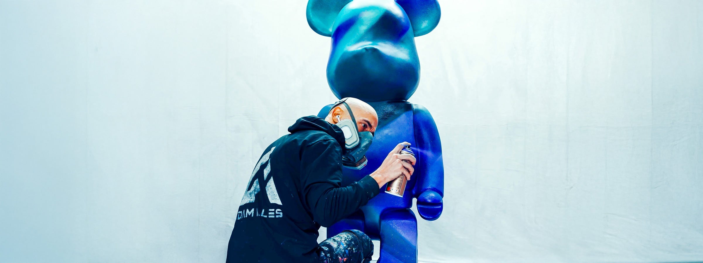 Artist Adam Illes wearing a respirator mask and black hoodie spray-paints a large blue bear sculpture in shades of blue against a light backdrop.