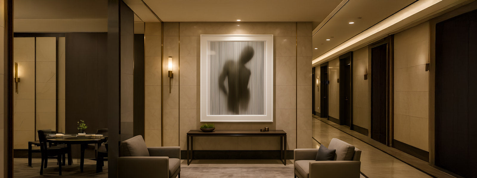 Framed art of a shadowy human silhouette behind vertical lines on a marble wall above a dark console table in a lobby with grey armchairs.