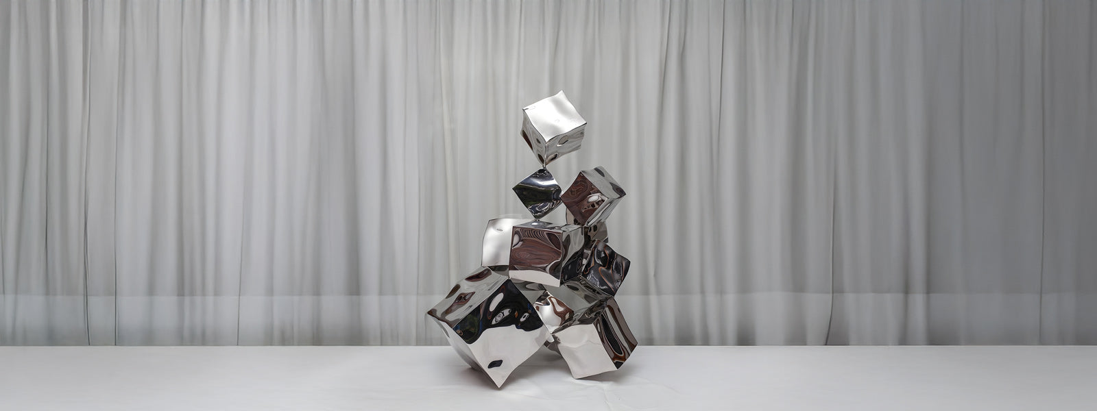Polished stainless steel sculpture of crushed and stacked cube forms in a dynamic balancing arrangement on a white surface with a white curtain backdrop.