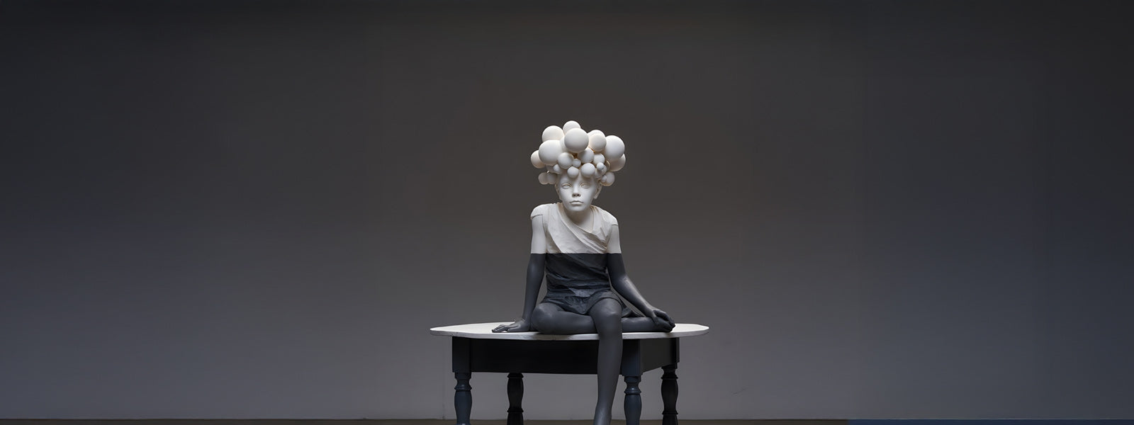 A sculpture of a seated child on a round table features a surreal design, with a cluster of white spheres as hair and contrasting black and white colors on the figure’s body and clothing.