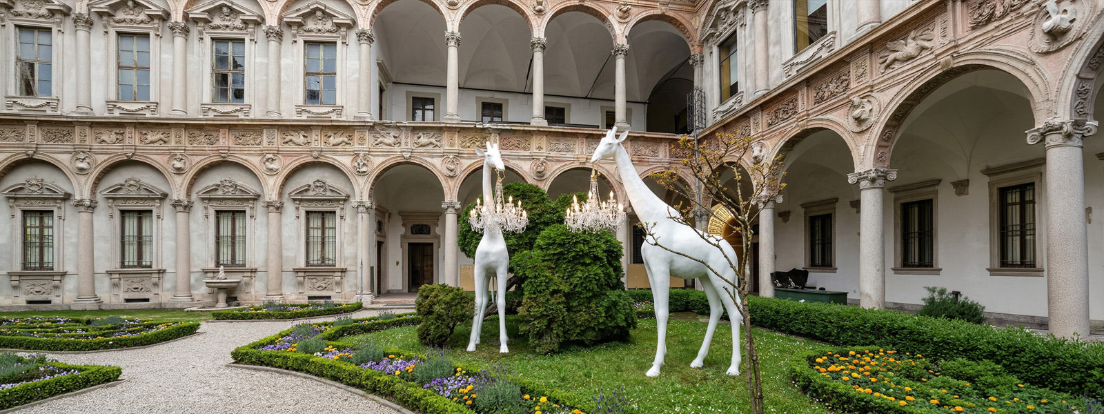 Two large white giraffe statues holding crystal chandeliers on a manicured courtyard lawn with hedges and flowers, framed by classical arched architecture.