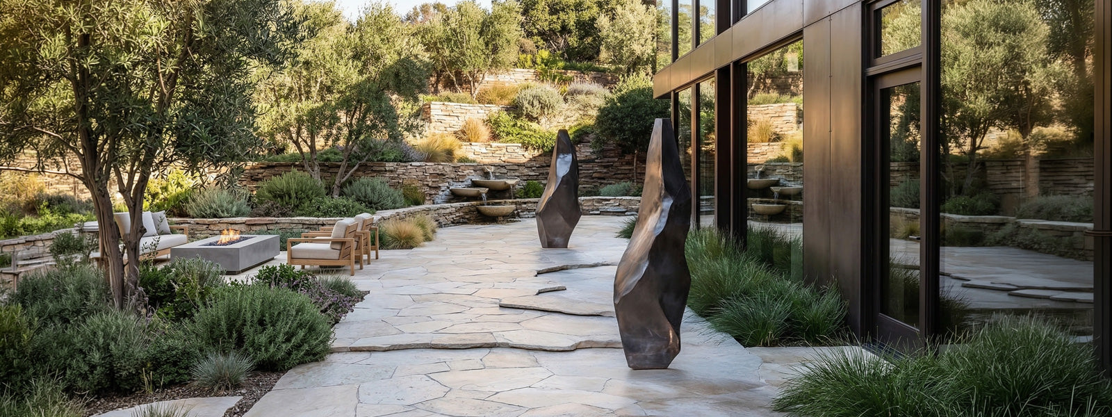 Two tall angular organic sculptures with dark bronze finish on a flagstone patio beside a modern glass building, flanked by olive trees and grasses.