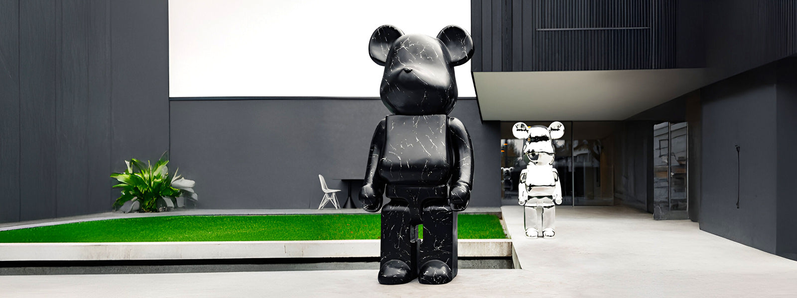 Large black marble-finish bear statue in a modern grey courtyard with green lawn, white chair, and smaller white marble-finish bear near a glass entrance.