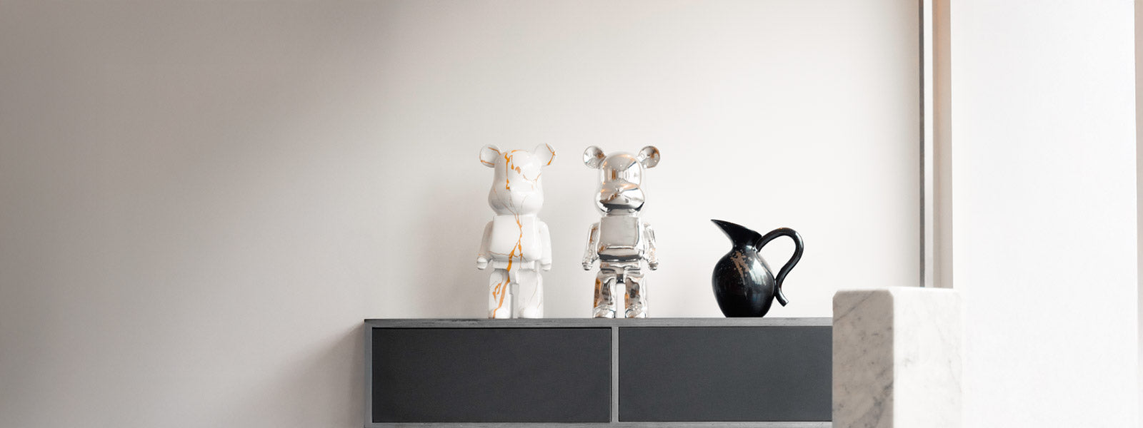 Two small bear sculptures, one white with gold-vein detailing and one in reflective chrome, on a dark grey sideboard beside a black ceramic pitcher.