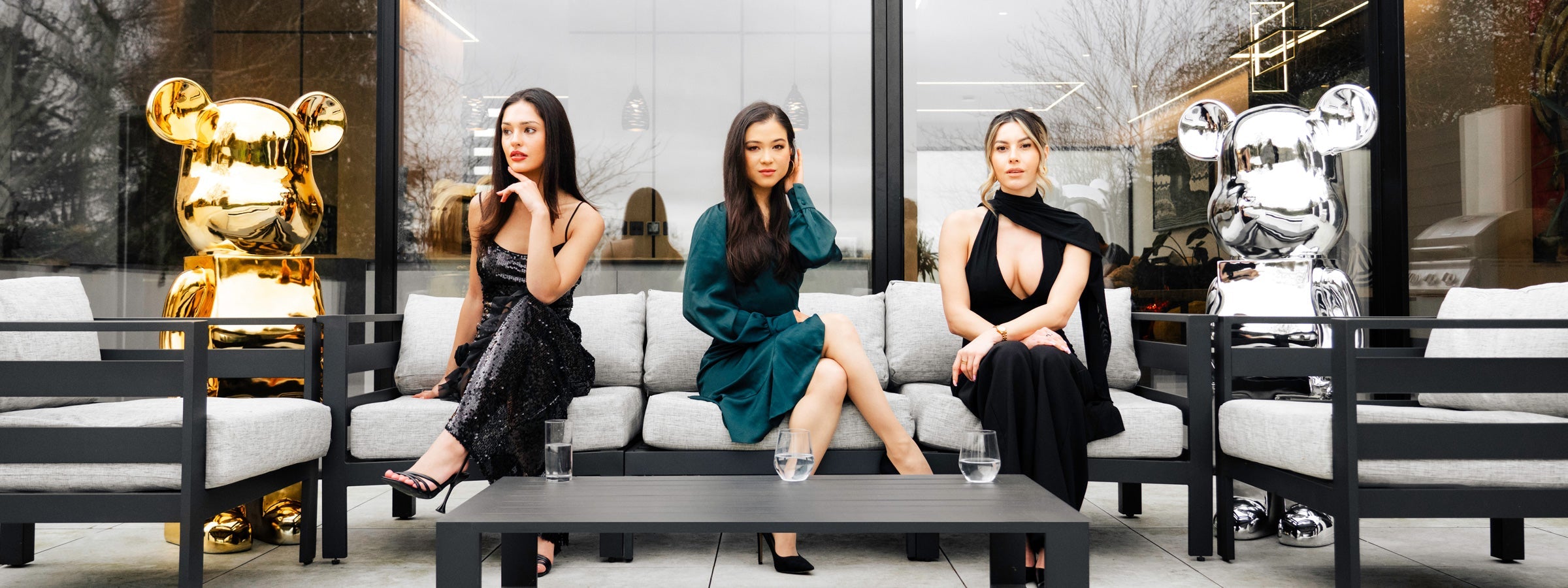 Three women in evening dresses seated on a grey outdoor sofa between a gold and a chrome medium bear sculpture with a modern glass-walled interior behind.
