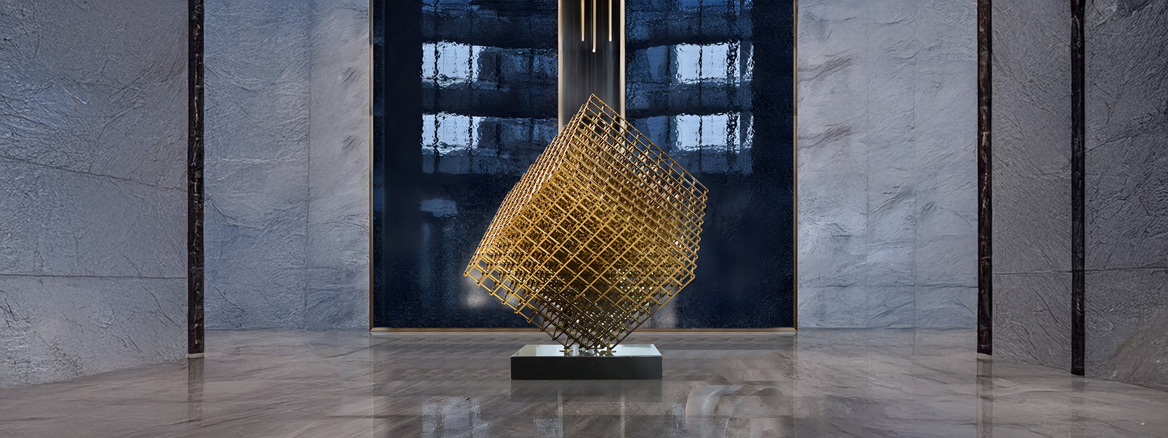 Gold geometric cube sculpture of intersecting grid bars balanced on one corner on a black pedestal in a modern lobby with stone walls and marble floor.