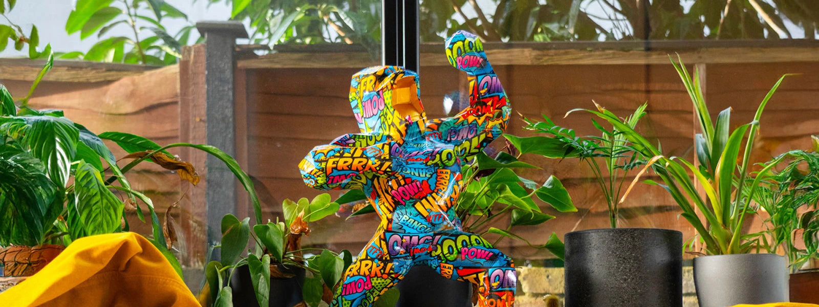 Pop art gorilla sculpture covered in colorful comic-book words like POW, BANG, OMG, and FRRR, displayed among potted green plants by a glass window.