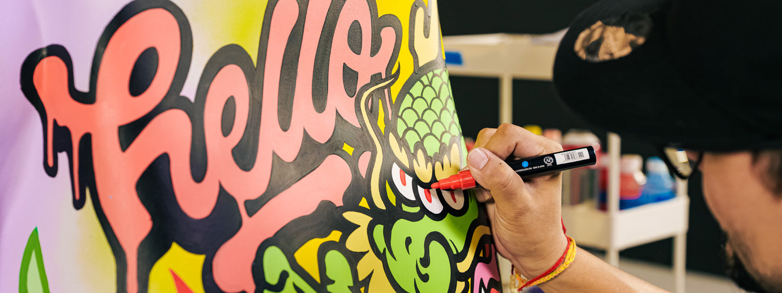 Artist in a black cap hand-details a colorful graffiti-style painting with bold pink "Hello" lettering and a green dragon character using a red-tipped marker.