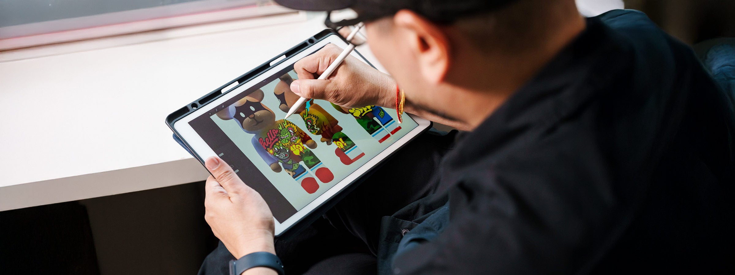 A person using a stylus to draw colorful cartoon characters on a tablet, seated by a window.