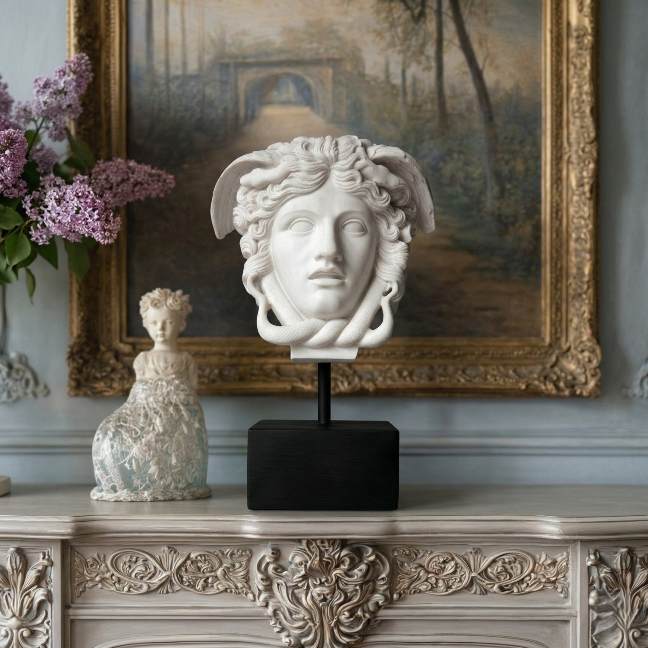 The Medusa Head Marble Sculpture - 80cm by Giant Sculptures, with wings and set on a black stand, is displayed atop an ornate mantel with a framed painting, porcelain figurine, and lilac flowers accenting this elegant marble arrangement.