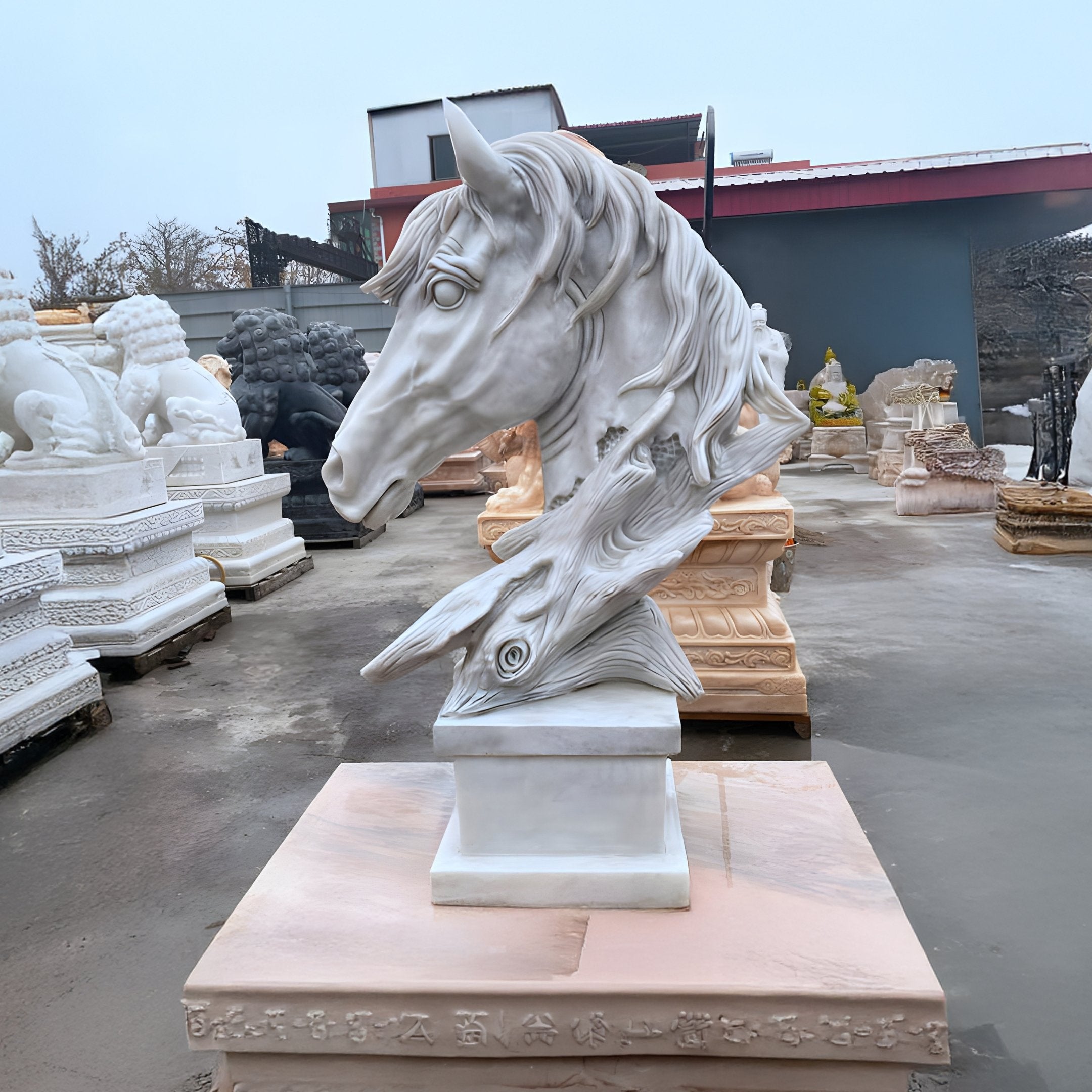 The Dynamic Horse Head White Marble Outdoor Sculpture (80cm) by Giant Sculptures features a horses head merging into a bird. Serving as an outdoor centerpiece on a pedestal with surrounding stone works, its striking presence is enhanced by an overcast sky and gray concrete ground.