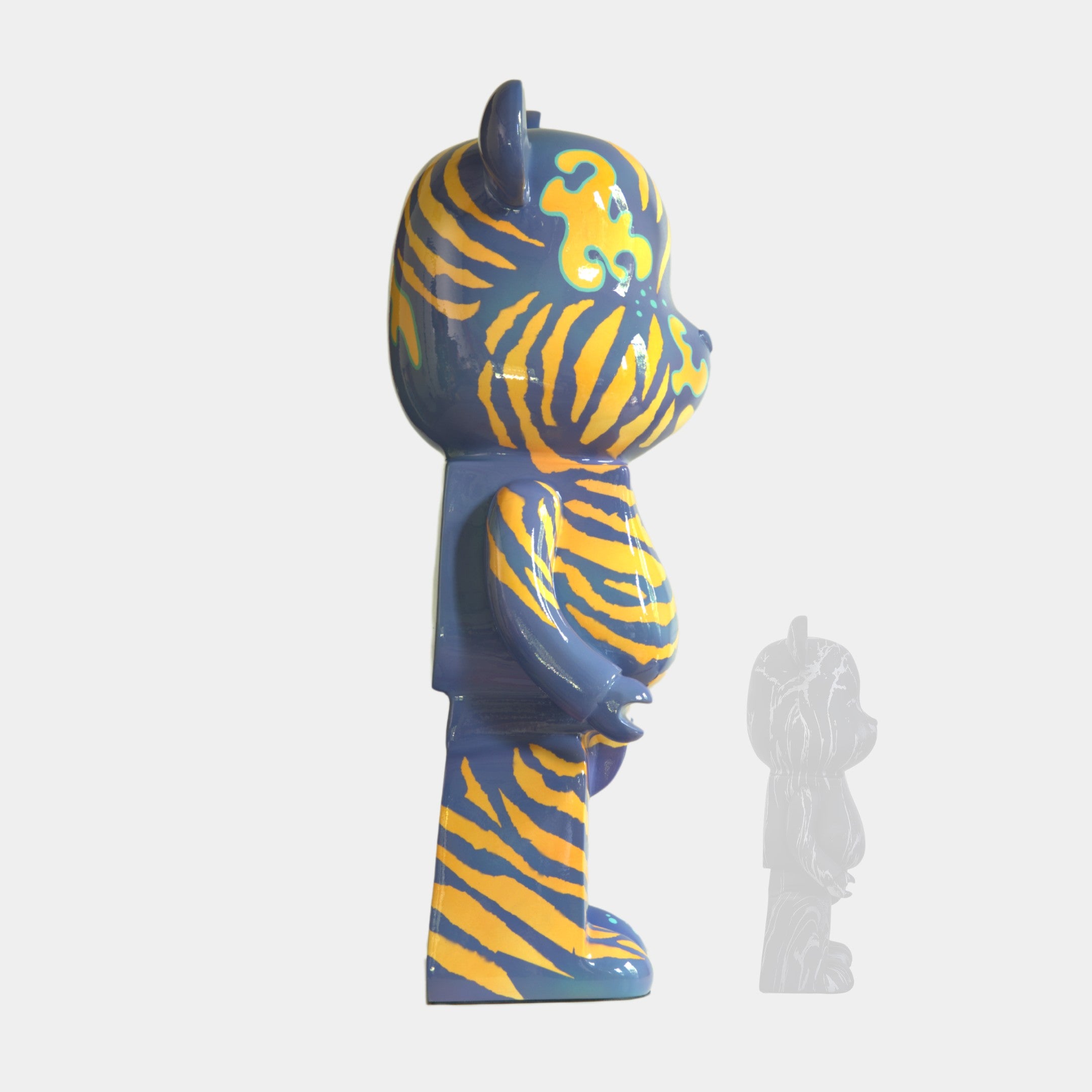 A side view of the Inside Appears Outside 2 Bear Sculpture - 50cm by Giant Sculptures, featuring blue and yellow tiger stripes, stands upright on a white background with a faded, smaller version visible behind it.
