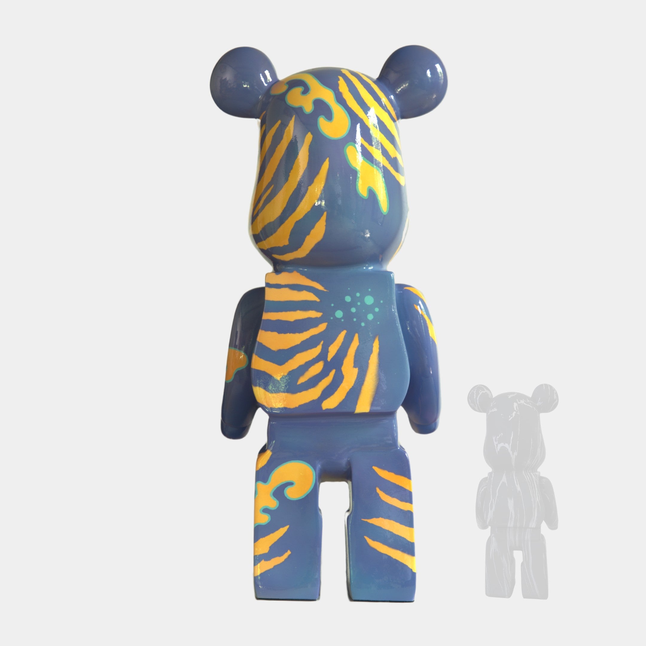 The Inside Appears Outside 2 Bear Sculpture (50cm) by Giant Sculptures is a blue, bear-shaped vinyl toy with yellow and orange wavy stripes and green spots, facing backward. A faint gray outline appears to its right on a plain white background.