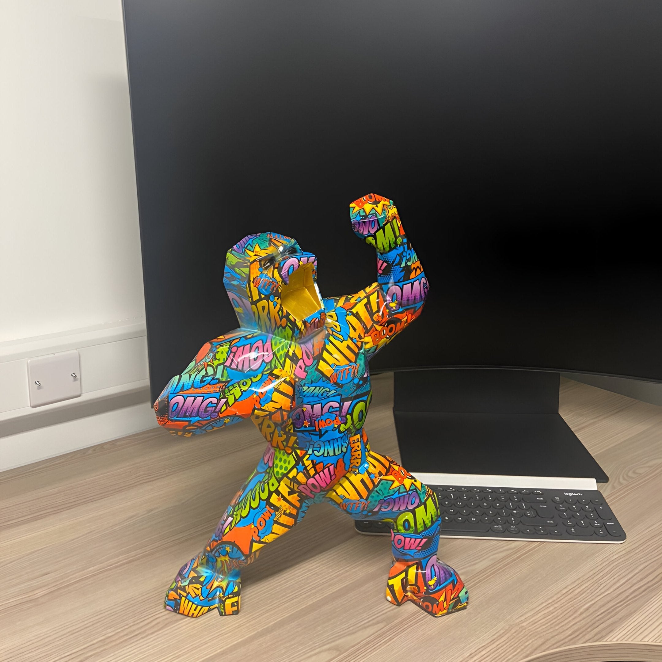 The Giant Sculptures Pop Art Gorilla Sculpture - 50cm (Ex Display) features comic book-style words like WOW and OMG, flexing boldly on a desk to add striking contemporary decor in front of your computer monitor and keyboard.
