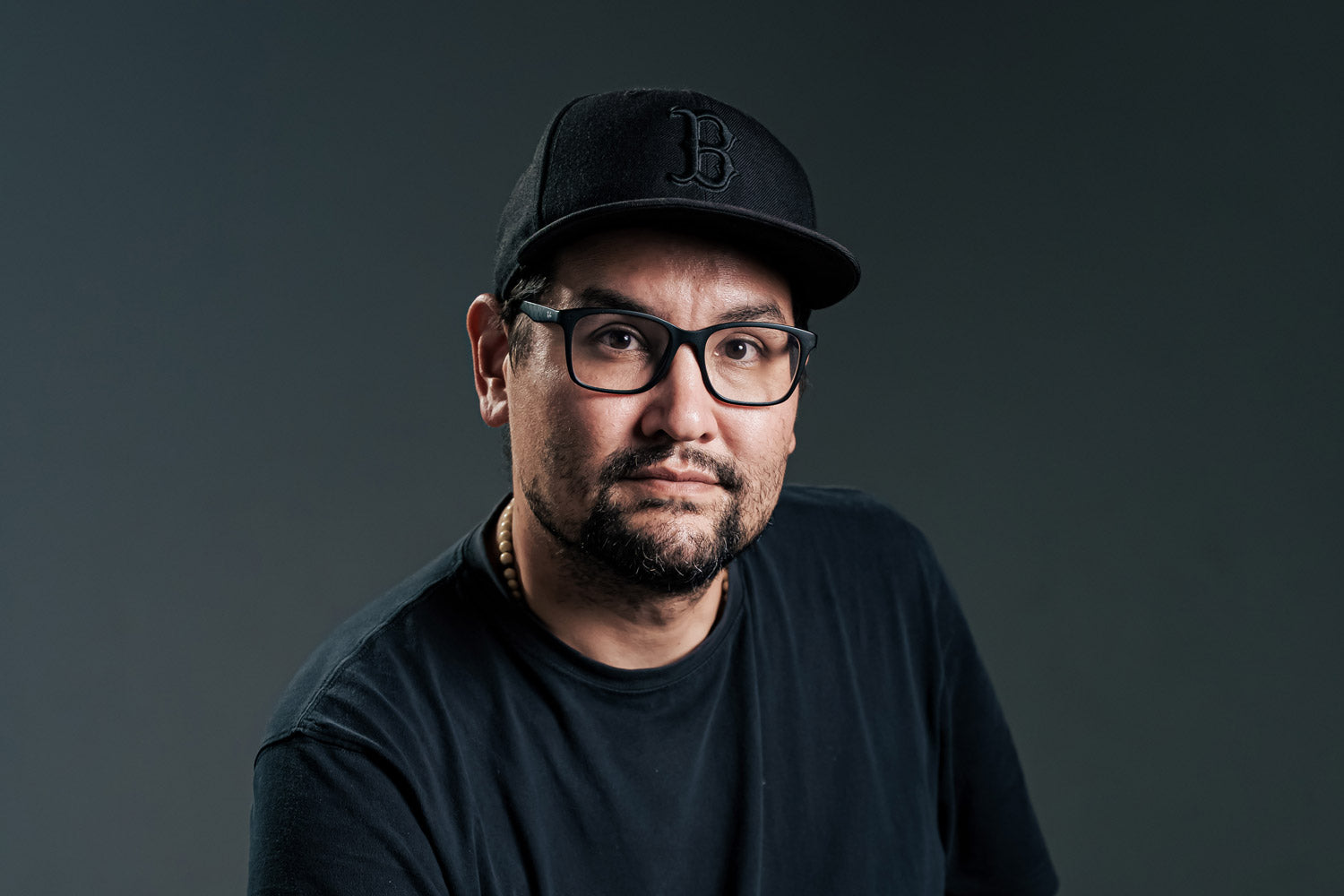A man with a beard and mustache, wearing black glasses, a black baseball cap with a B logo, and a black t-shirt, poses against a dark gray background.