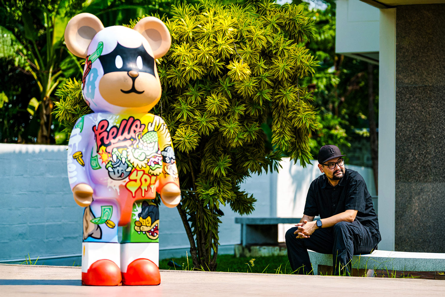 A man in black clothing and hat sits on a bench outdoors, looking at a large, colorful bear statue decorated with graffiti-style art and the word hello. Greenery and a building are visible in the background.