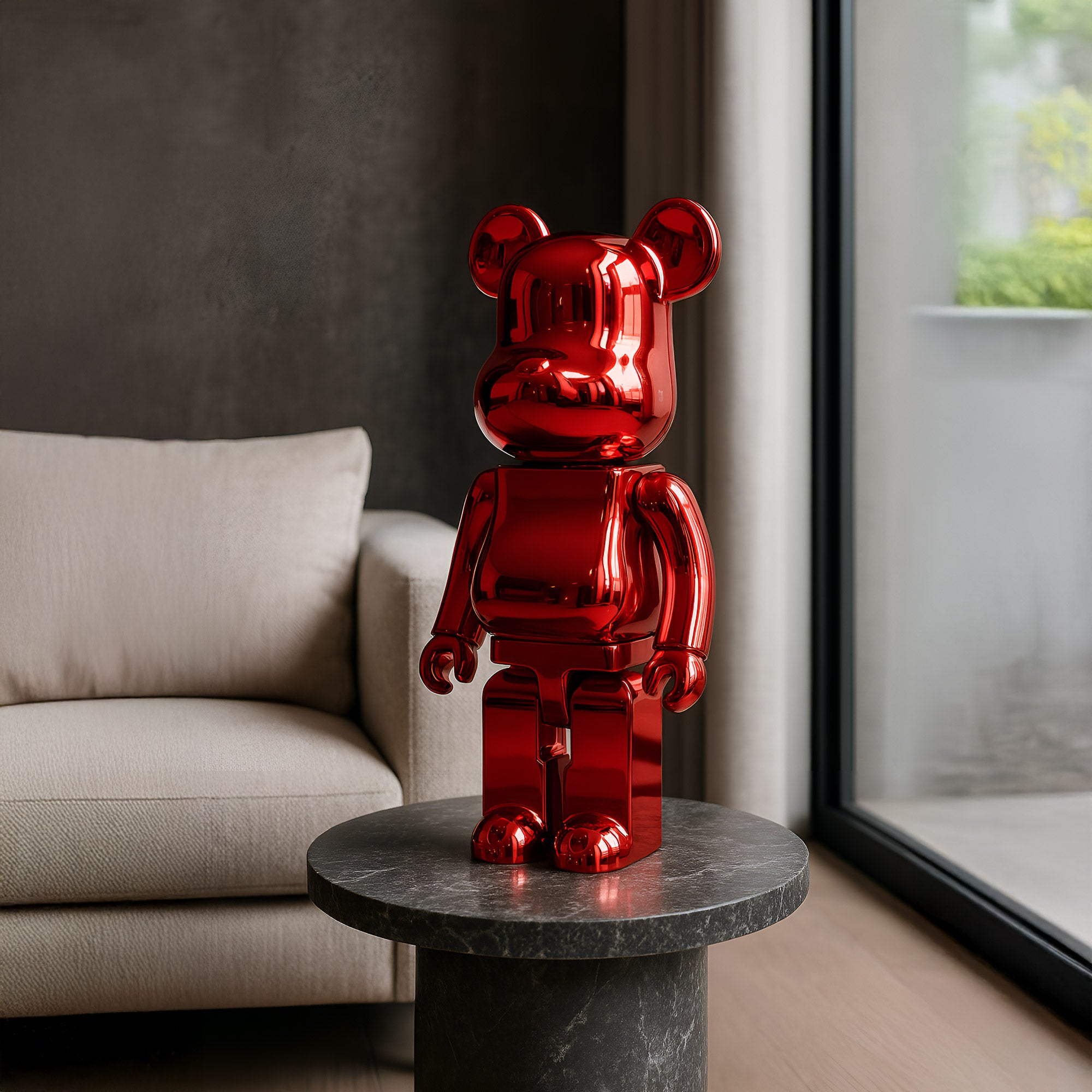 The 50cm Ruby Red Bear Sculpture by Giant Sculptures, featuring an electroplated finish, stands on a round gray marble table in a modern living room. Next to a beige sofa and large windows showcasing outside greenery, it embodies luxurious artistry.