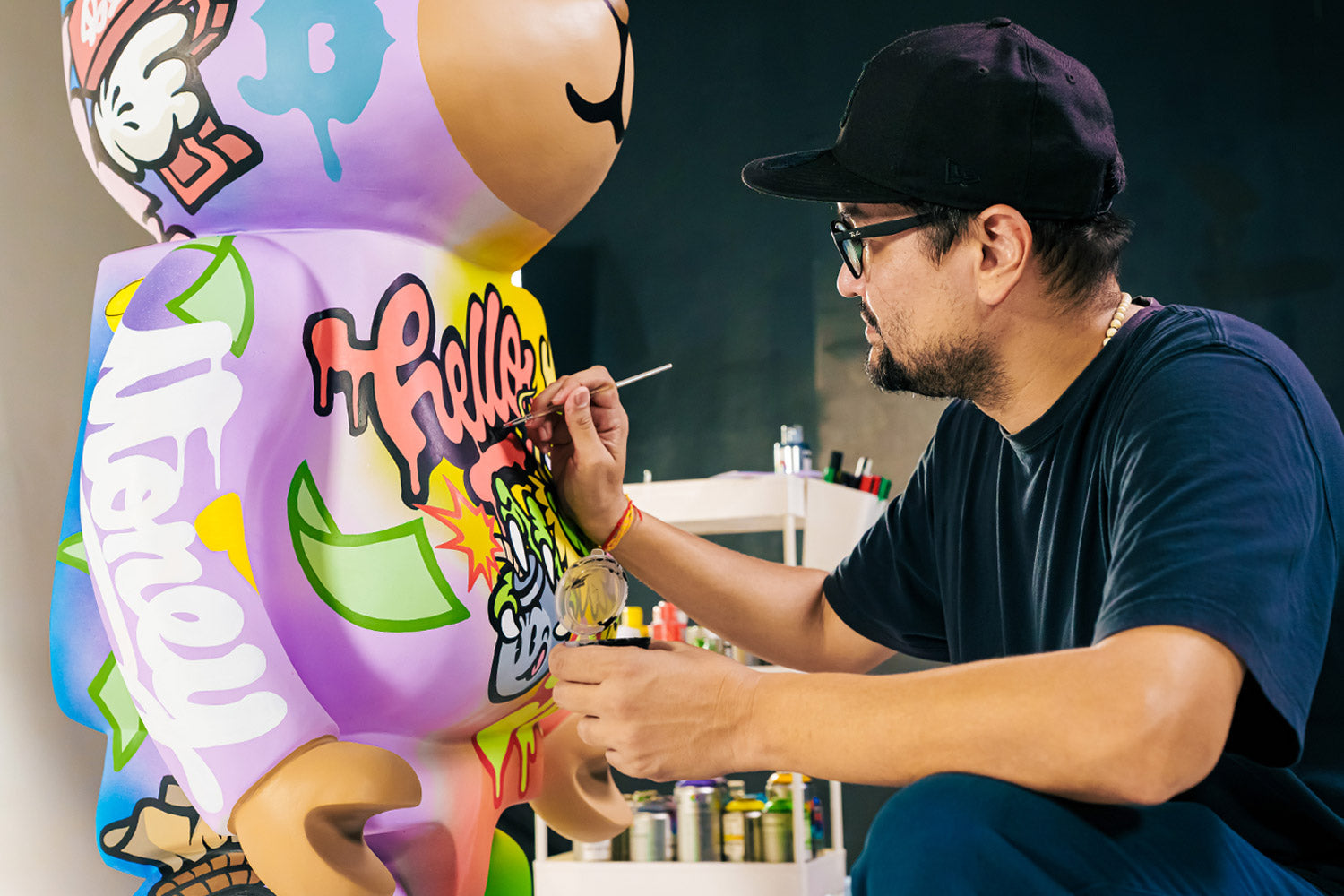 A man wearing glasses and a cap is painting colorful graffiti art on a large bear-shaped sculpture. He uses a fine brush, and the sculpture features vibrant words and cartoon-like designs. Art supplies are visible in the background.