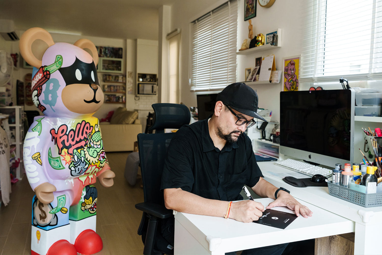 A man wearing glasses and a cap sits at a white desk, drawing on paper in a bright, art-filled studio. Beside him stands a large, colorful bear sculpture decorated with vibrant graffiti-style artwork.