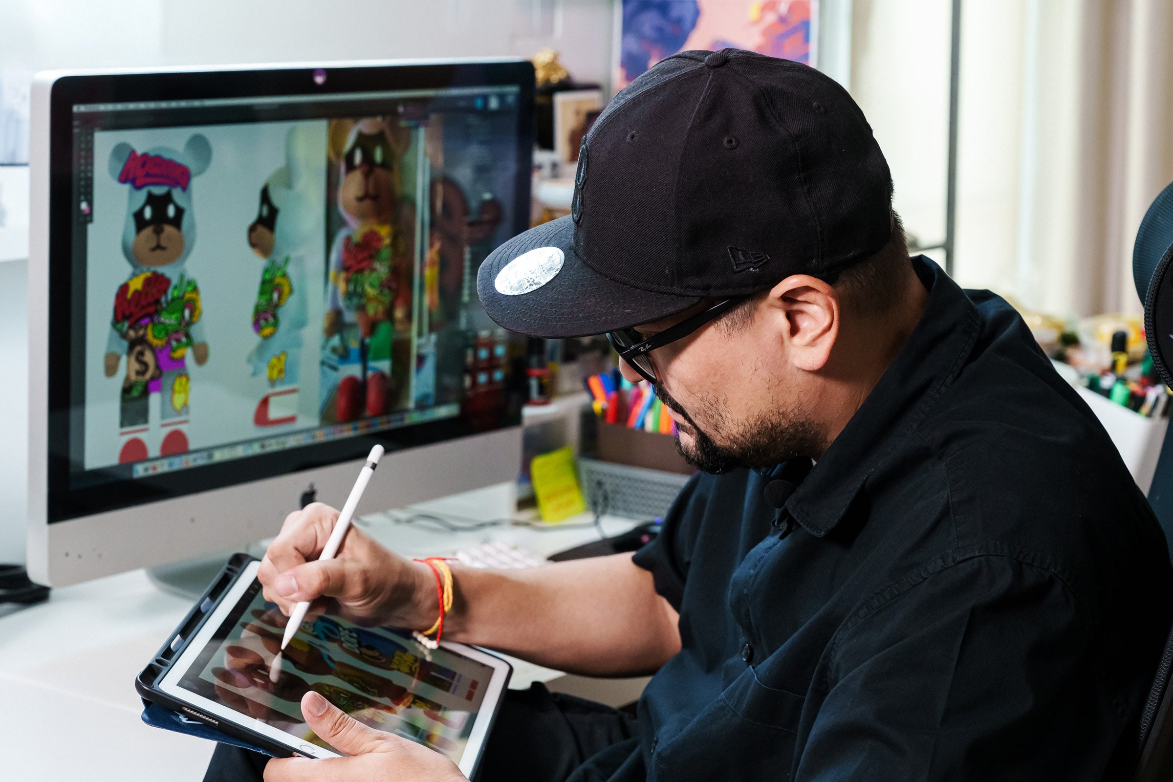 A person wearing a black cap and glasses draws colorful cartoon characters on a tablet, with similar artwork displayed on a large computer screen in the background.