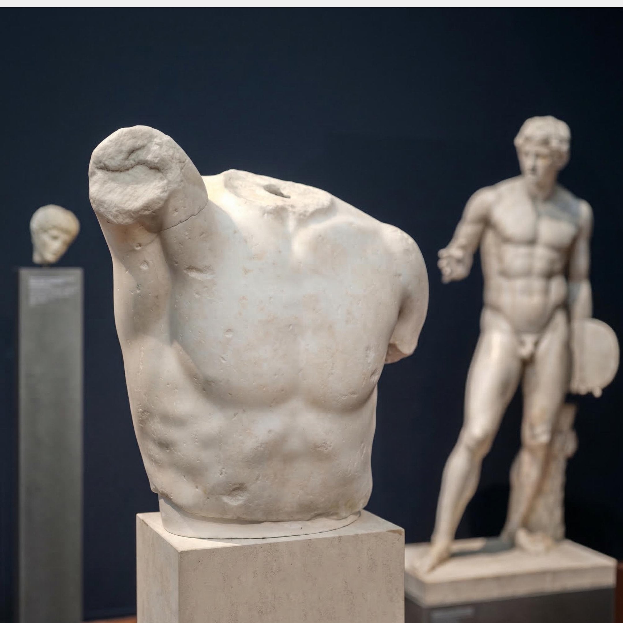 The Giant Sculptures Classical Male Torso Fragment Sculpture (80cm) stands headless and armless in the foreground, with figurative art—a full-body statue and a smaller sculpted head—displayed behind it in the museum.