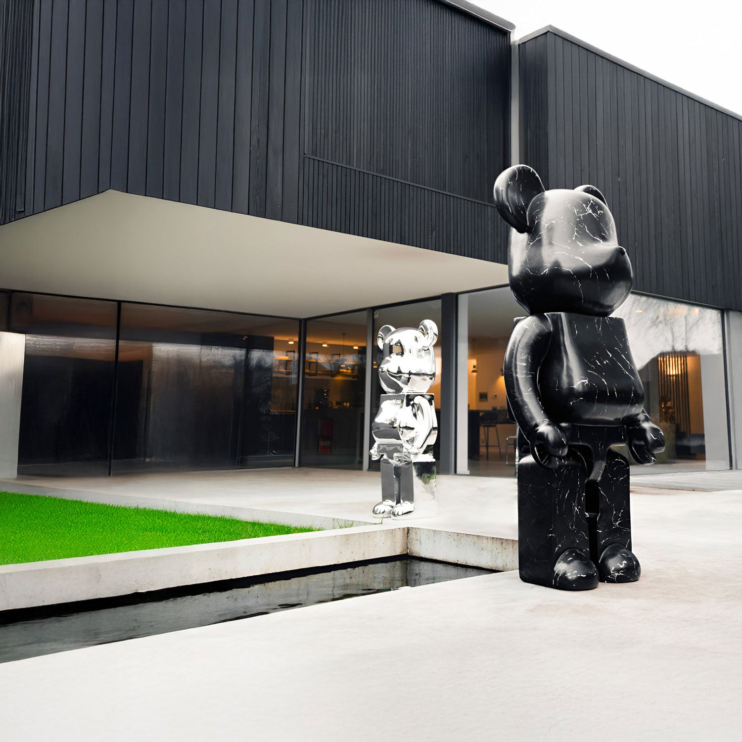 Two large bear sculptures, one black and one silver, stand outside a modern building with dark panels and large windows. The black sculpture is closer, set near a grassy rectangular patch and a narrow water feature.