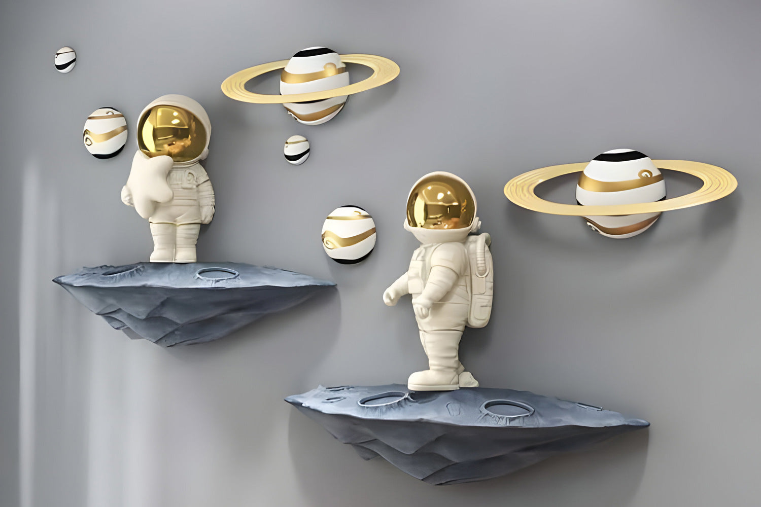Two astronaut sculptures with gold visors stand on rocky platforms. They are surrounded by small planets with rings, mounted on a grey wall. The scene resembles a decorative space-themed installation.