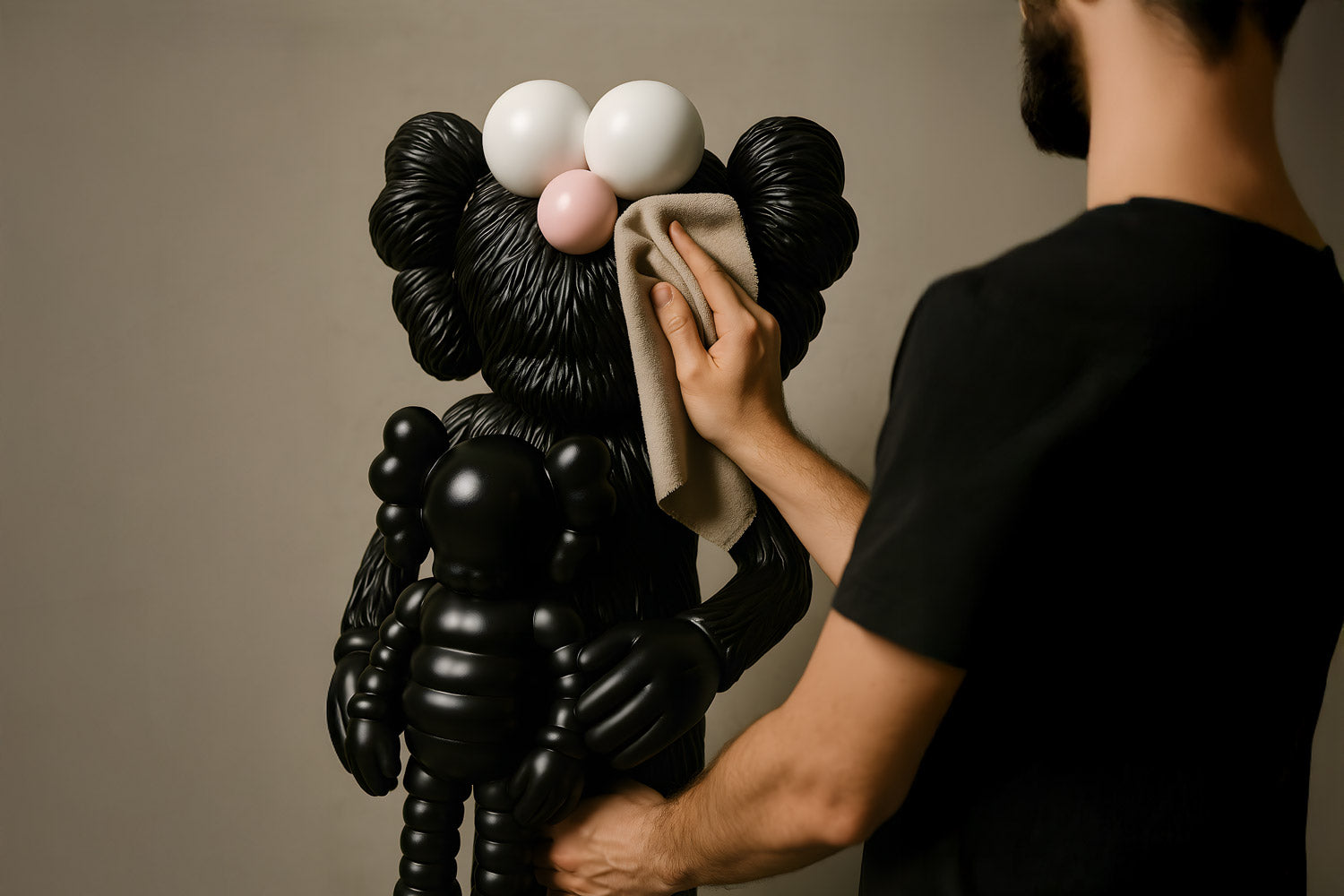 A person in a black shirt wipes a large black sculpted figure resembling a cartoon character with big eyes and a pink nose; the figure holds a smaller similar figure.