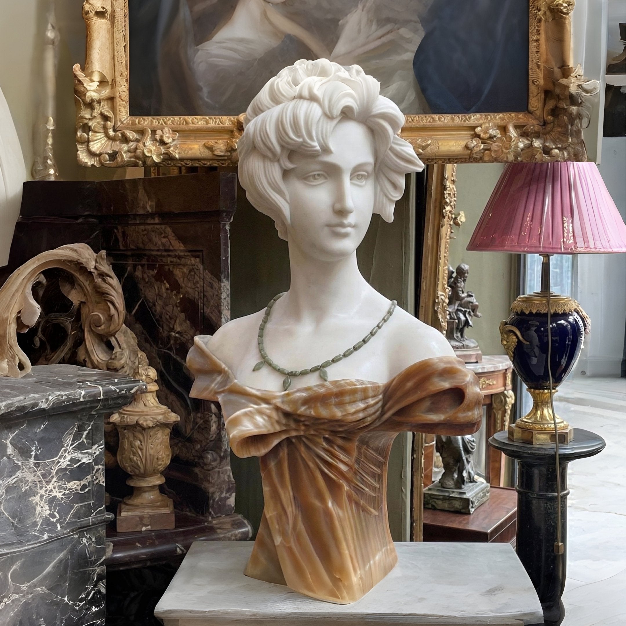 The Elegant Lady Marble Bust Sculpture - 60cm by Giant Sculptures features a wavy-haired lady with a necklace, displayed indoors among ornate furniture, a framed painting, a pink lampshade, and decorative objects.