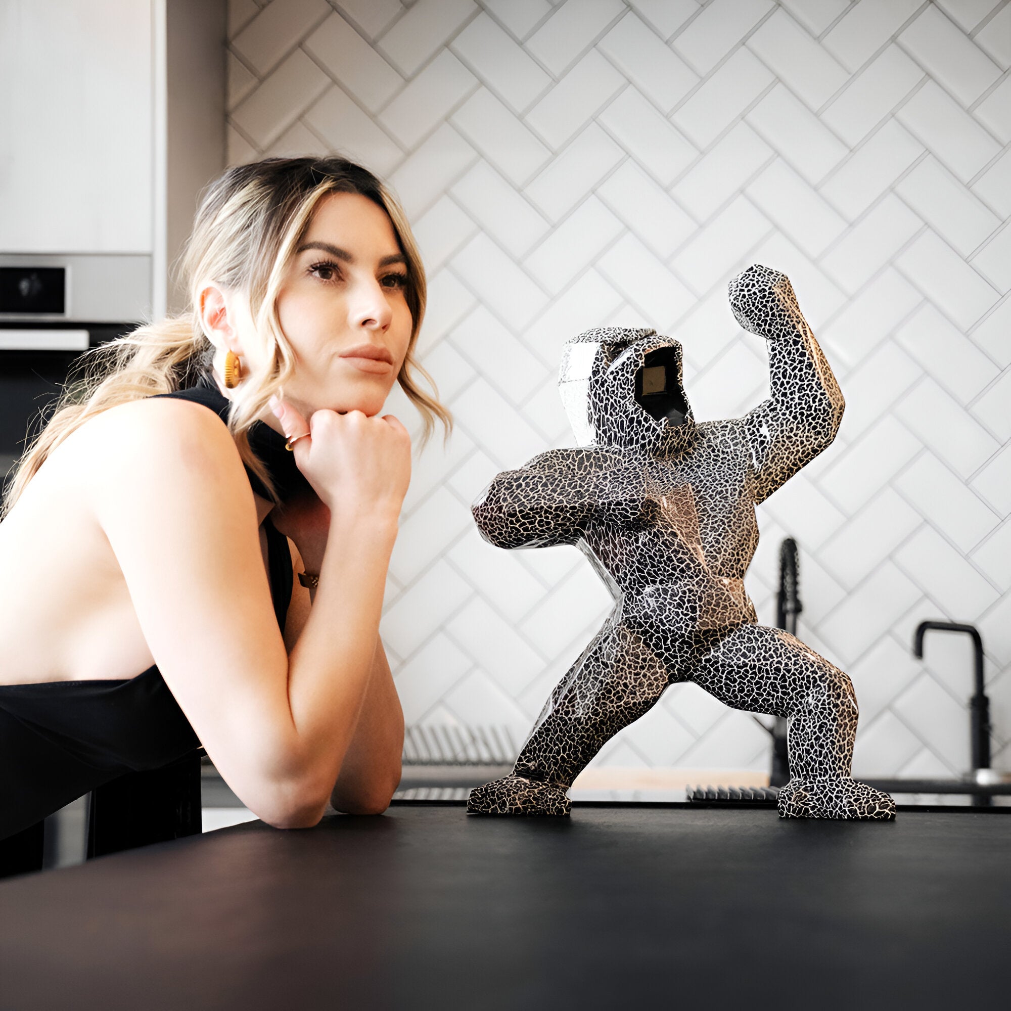 A faceted black and white textured gorilla sculpture with a cracked pattern, positioned on a kitchen counter with a woman in the background, looking contemplative.