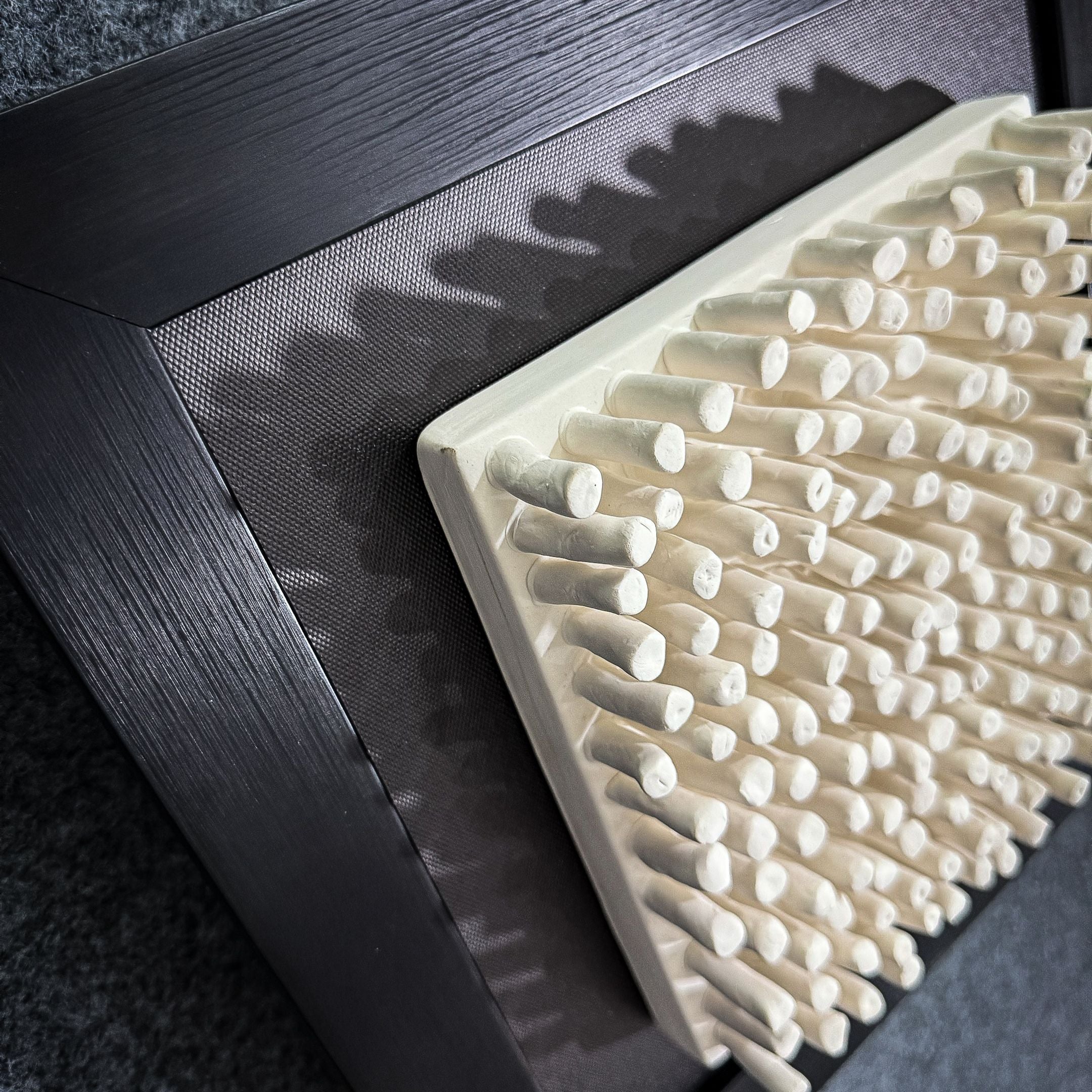 Close-up of the Harmonia 3D Ceramic Grid Wall Sculpture With Black Frame by Giant Sculptures—white cylindrical pegs on a rectangular board, angled in a dark frame—adds striking contemporary style to modern interiors.