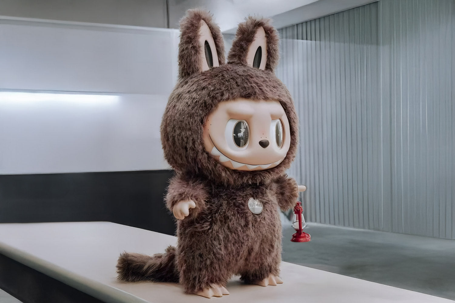 A large, Labubu figure in a fluffy brown bunny costume with big ears, sharp teeth, and wide eyes stands indoors holding a small red plunger. The background is modern and minimalistic.