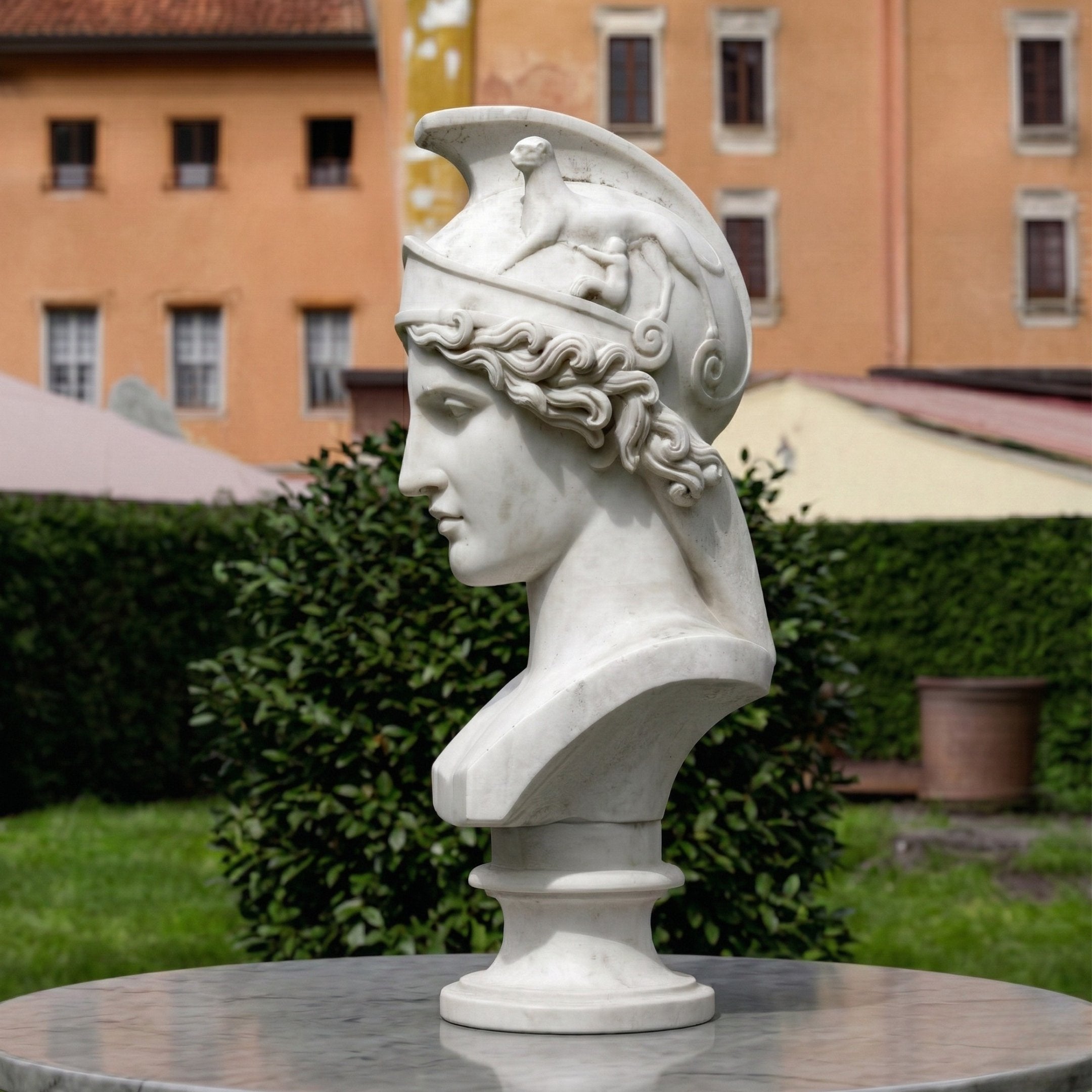 The Athena Marble Bust Sculpture - 65cm by Giant Sculptures depicts a classical figure with a winged, horse-adorned helmet, displayed outdoors on a round marble pedestal amid green hedges and an orange building.