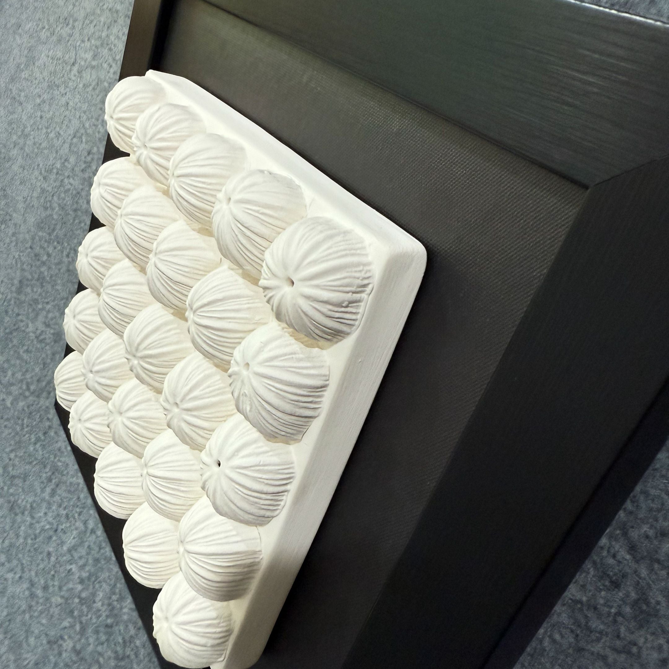 An angled view of the Harmonia 3D Floral Grid Sculpture With Black Frame by Giant Sculptures, featuring white floral-inspired ceramic forms in a black rectangular frame, mounted on a blue-gray textured wall.