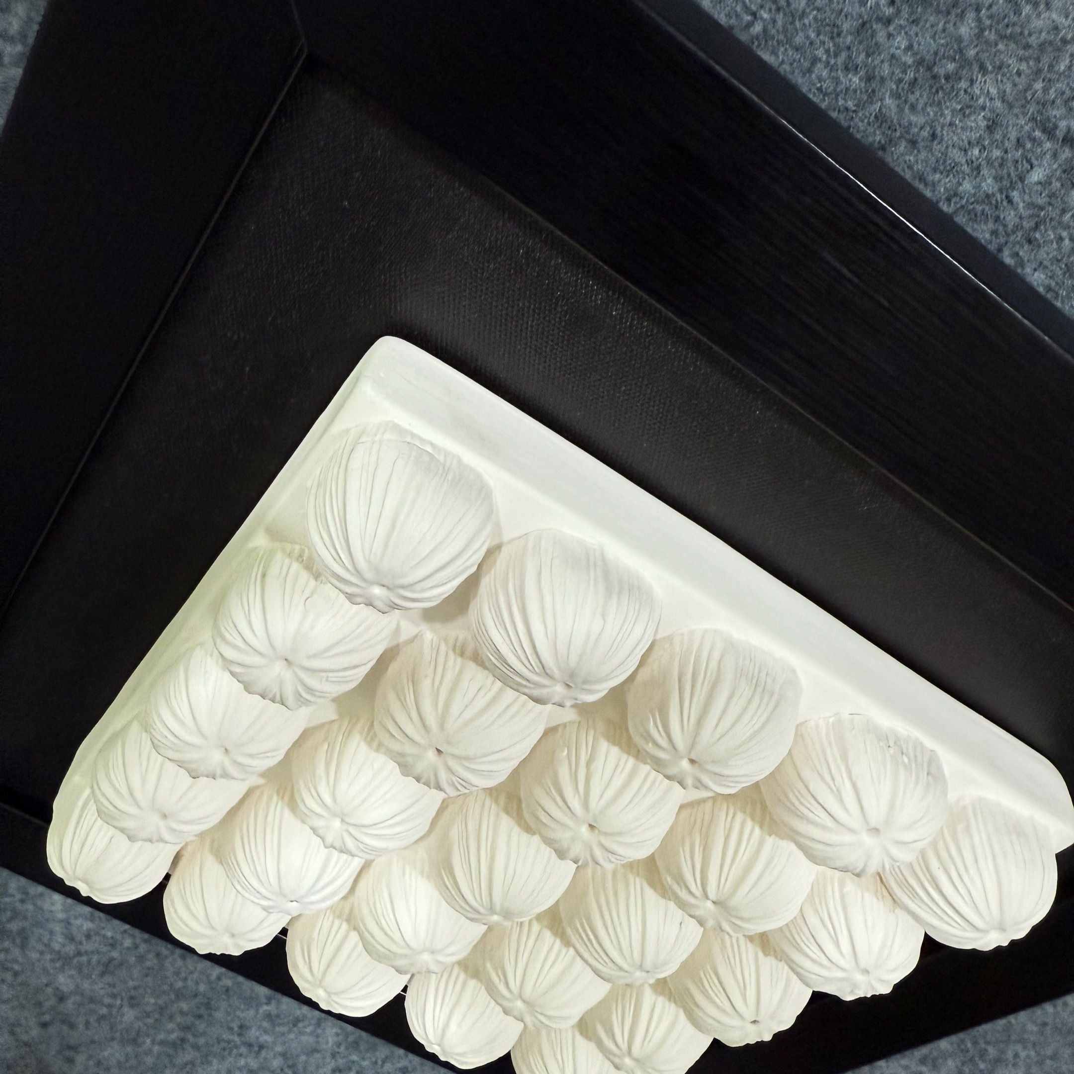 A close-up of the Harmonia 3D Floral Grid Sculpture by Giant Sculptures features white, shell-shaped bulbs in a grid within a black frame, mounted on a gray textured ceiling for modern charm.