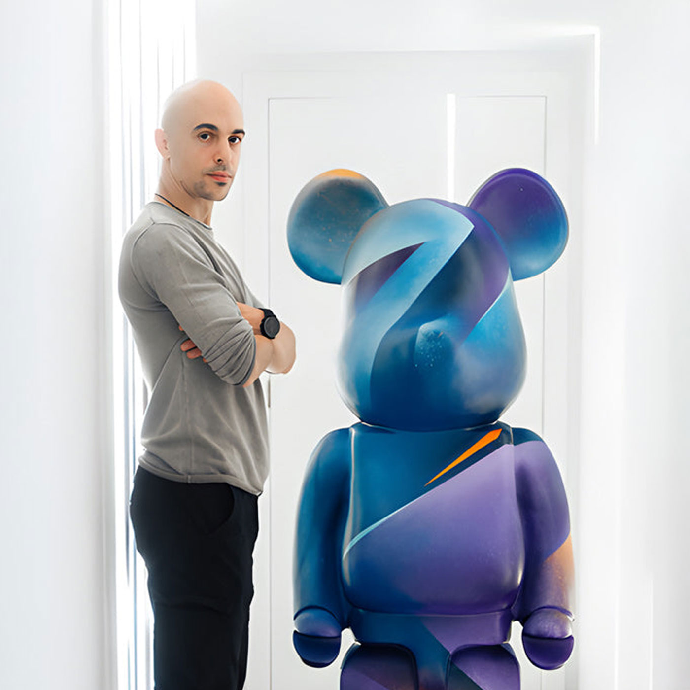 A bald man stands with arms crossed next to a large, colorful bear-shaped sculpture with abstract blue and purple patterns in a bright, modern room.