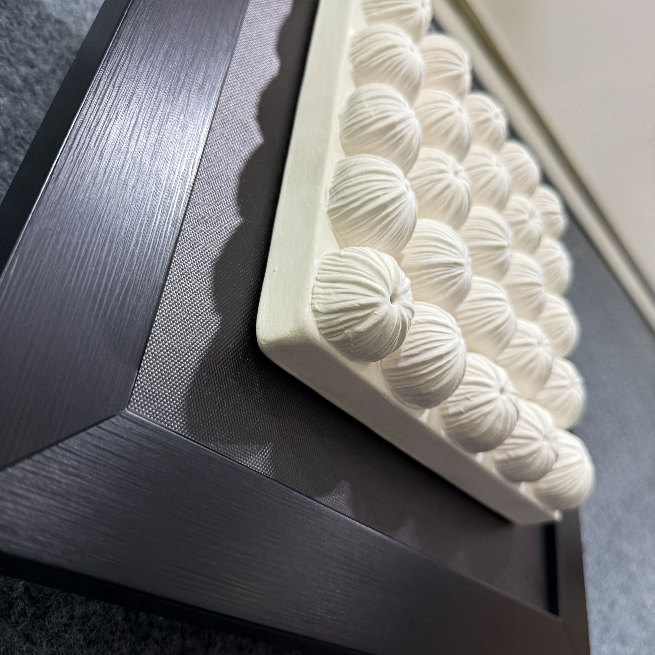 The Harmonia 3D Floral Grid Sculpture With Black Frame by Giant Sculptures features rows of round, floral-inspired forms on a white tray set atop a dark frame, creating a layered, contemporary geometric look.