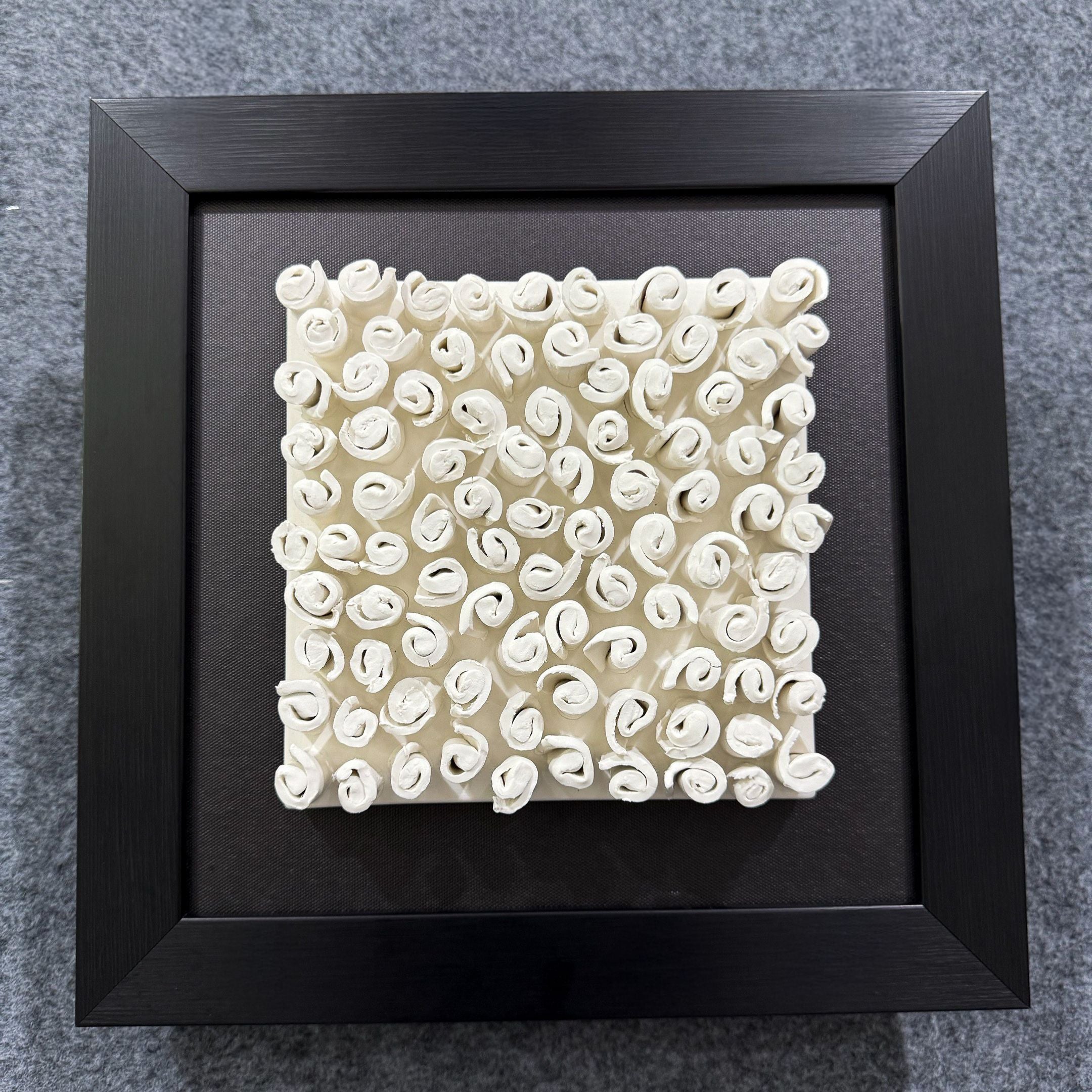 The Harmonia 3D Spiral Wall Sculpture With Black Frame by Giant Sculptures features small, white, rolled paper-like shapes arranged in a grid on black—an elegant square piece ideal for contemporary spaces.