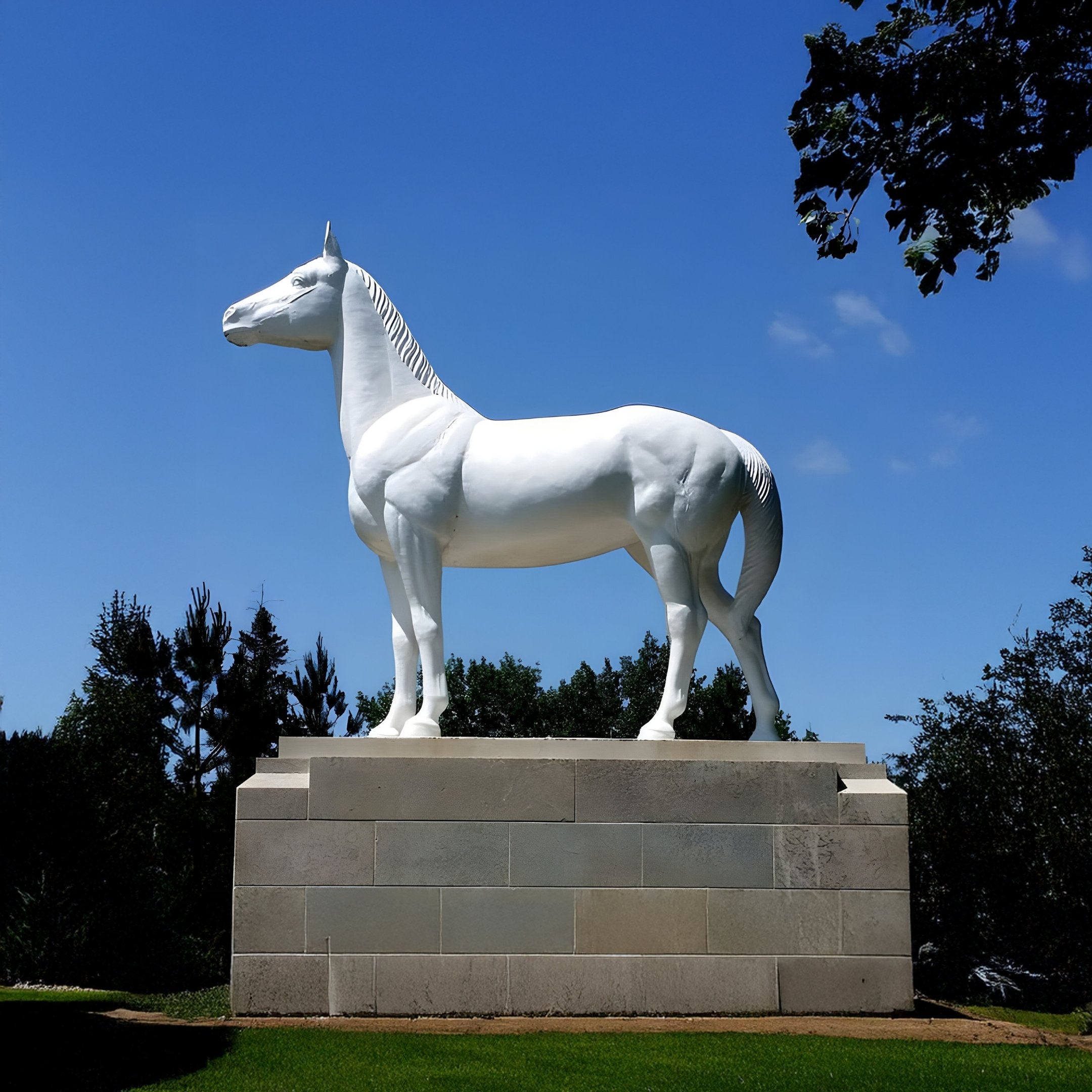 In a serene setting, the Noble Standing Horse Marble Outdoor Sculpture - 220cm by Giant Sculptures majestically stands on a stone pedestal against a clear blue sky, surrounded by green trees. Its hand-carved details in white marble capture an elegant scene.