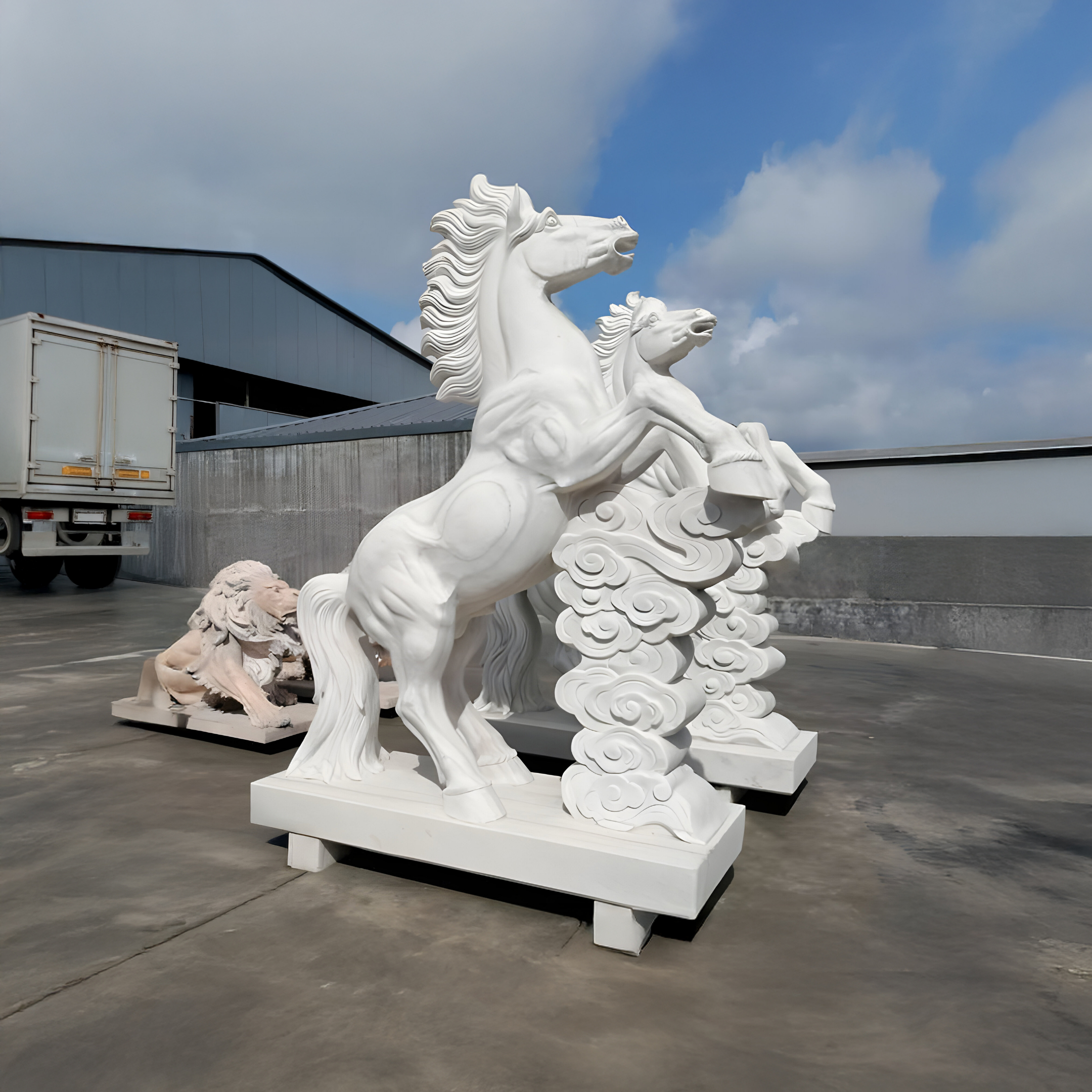 Two white marble horse statues, including the Cloud-Rearing Horse Marble Outdoor Sculpture by Giant Sculptures, stand on concrete. A crouching lion statue is behind them, while a truck and industrial building sit beneath a partly cloudy sky.