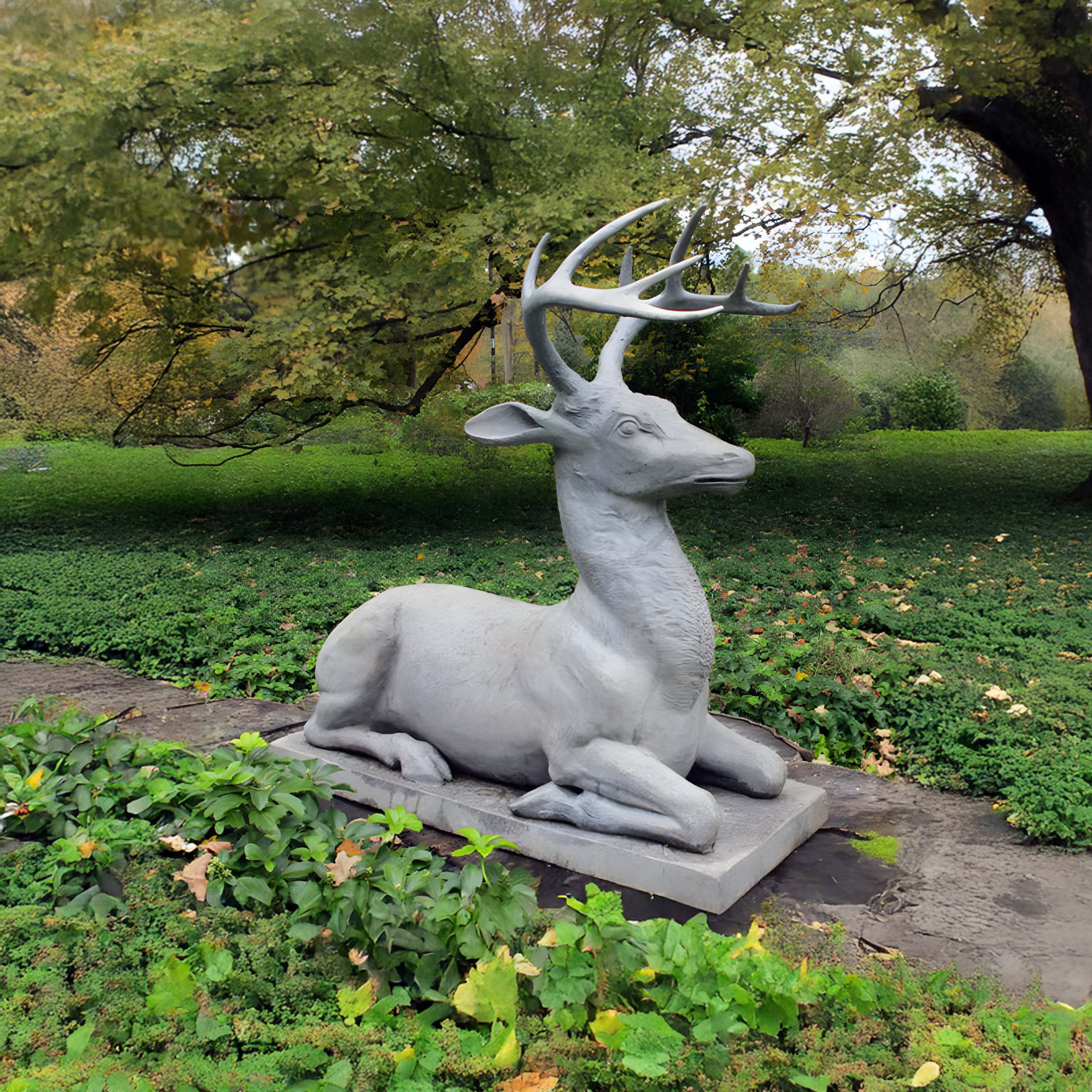 The Regal Lying Deer Marble Outdoor Sculpture - 180cm by Giant Sculptures depicts a gray deer with gracefully arched antlers on a rectangular base, perfectly complementing the lush grass and leafy trees of the park pathway.