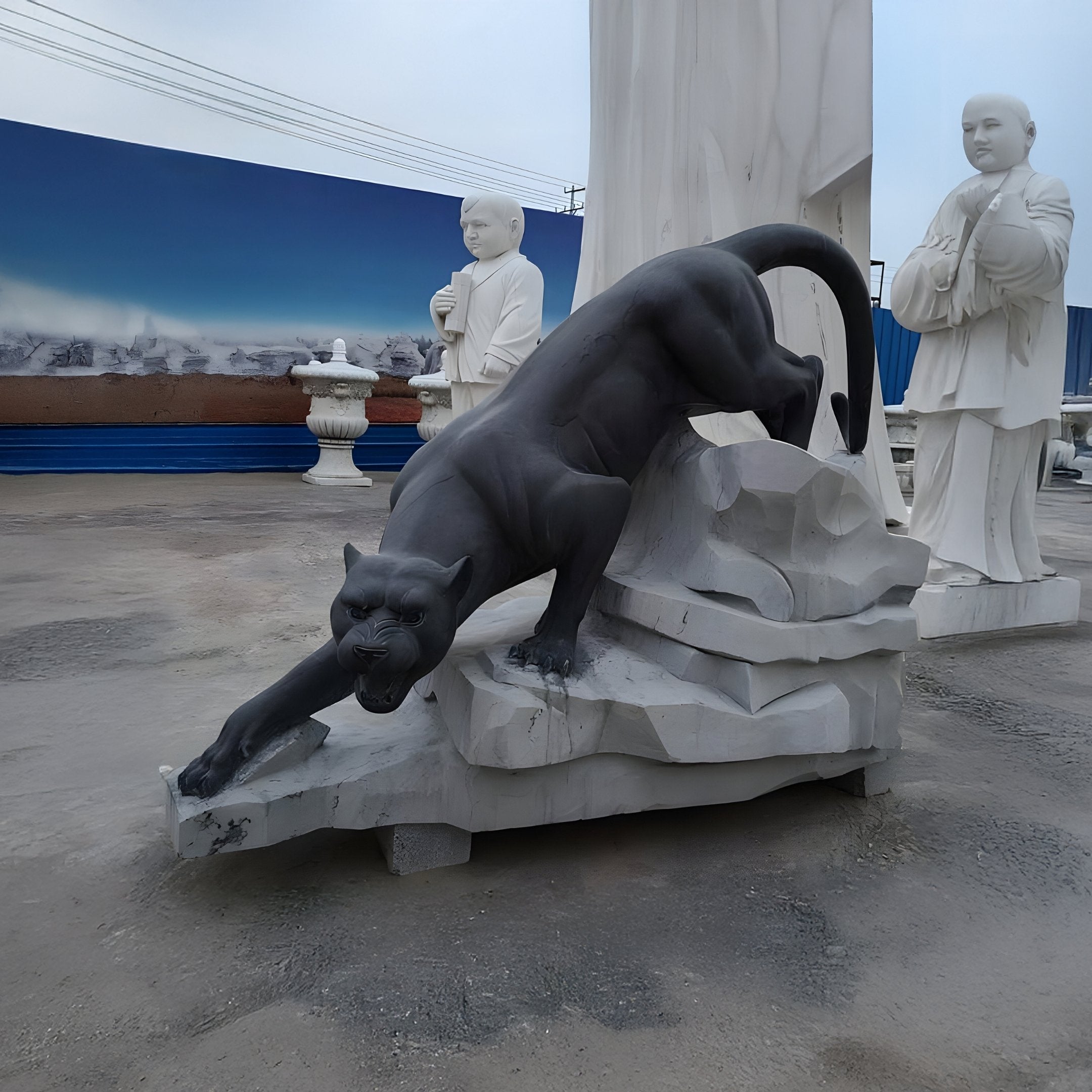 The Stealth Panther Marble Outdoor Sculpture by Giant Sculptures features a black panther poised to pounce on a rock-like base. Behind it, two white marble monks stand calmly against a serene gradient blue mural, suggesting an outdoor landscape.