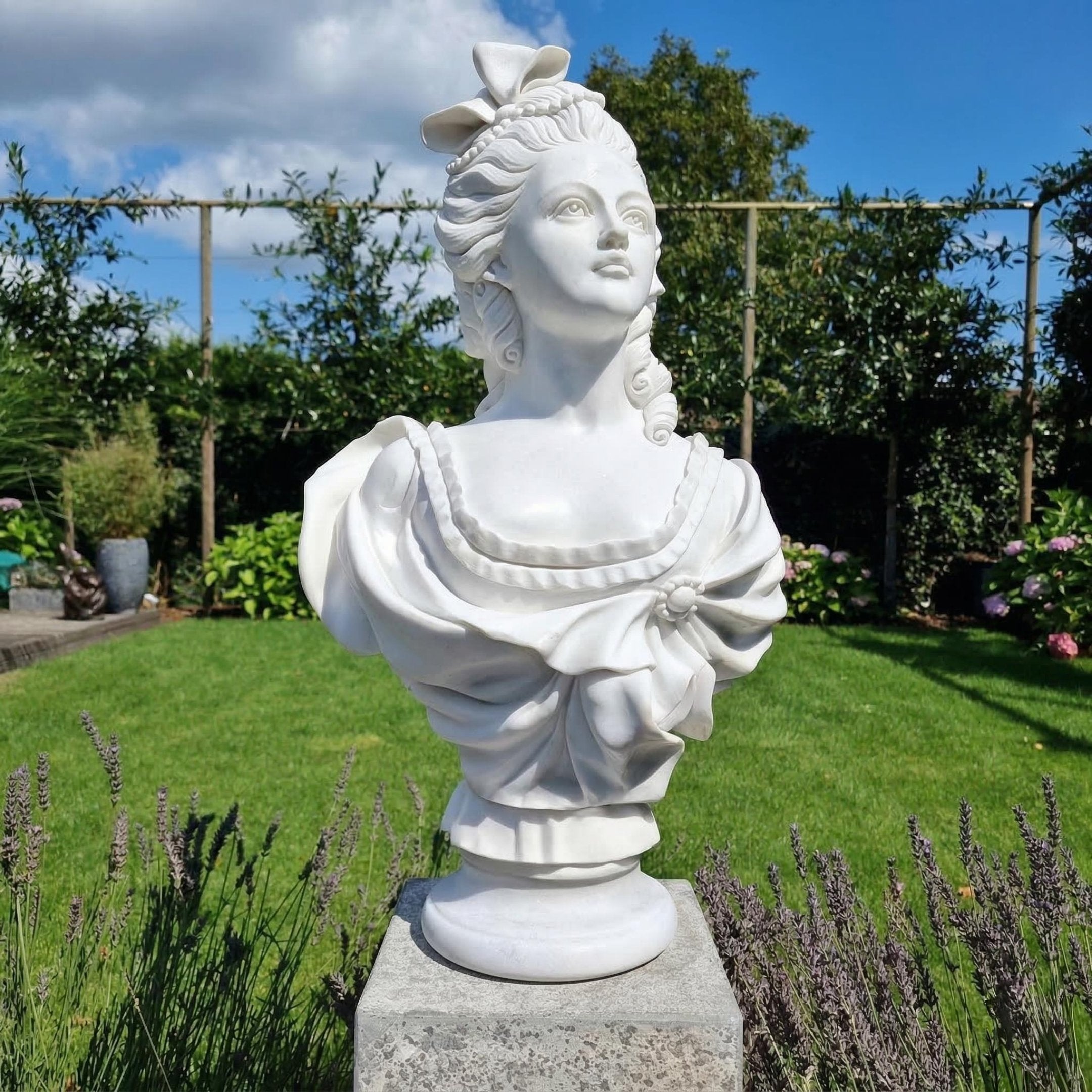 The Baroque Lady Marble Bust Sculpture - 65cm by Giant Sculptures, crafted in natural white marble with an elaborate hairstyle and draped dress, is displayed elegantly on a stone pedestal amid a lush garden of vibrant plants and flowers.