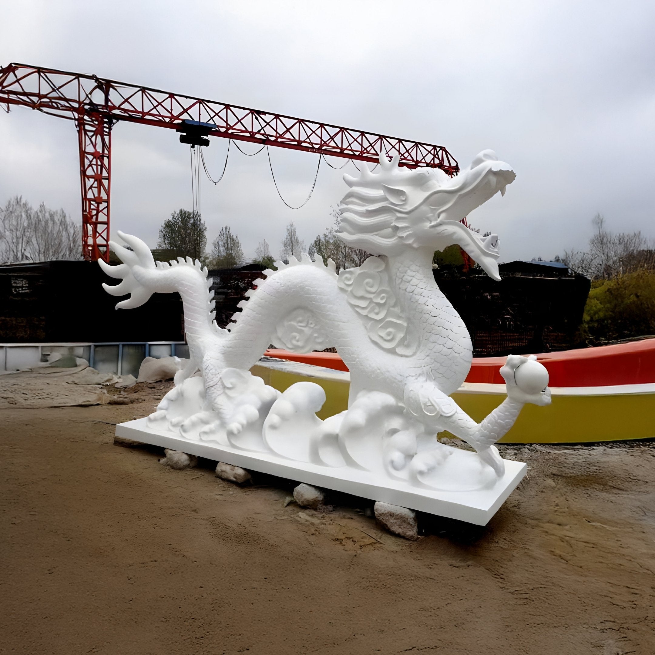 The Cloud-Surging Dragon Marble Outdoor Sculpture by Giant Sculptures, standing at 260cm, is a large, intricate white marble piece on a sandy surface. It looks energetic and dynamic with its open mouth and flowing form. A red metal structure and trees are seen in the background under a cloudy sky.