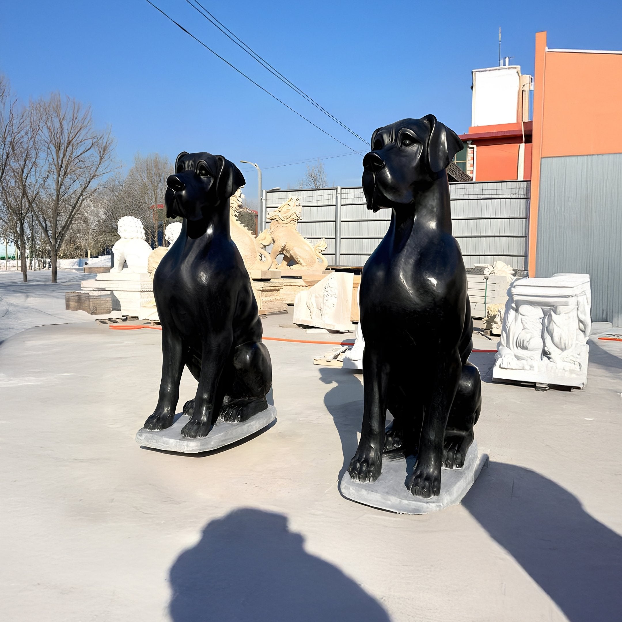 Two Ebony Guardian Dog Marble Outdoor Sculptures by Giant Sculptures, each 150cm tall, stand on concrete under a clear blue sky. Behind them are various sculptures like a lion and abstract shapes, with buildings and trees in the background.