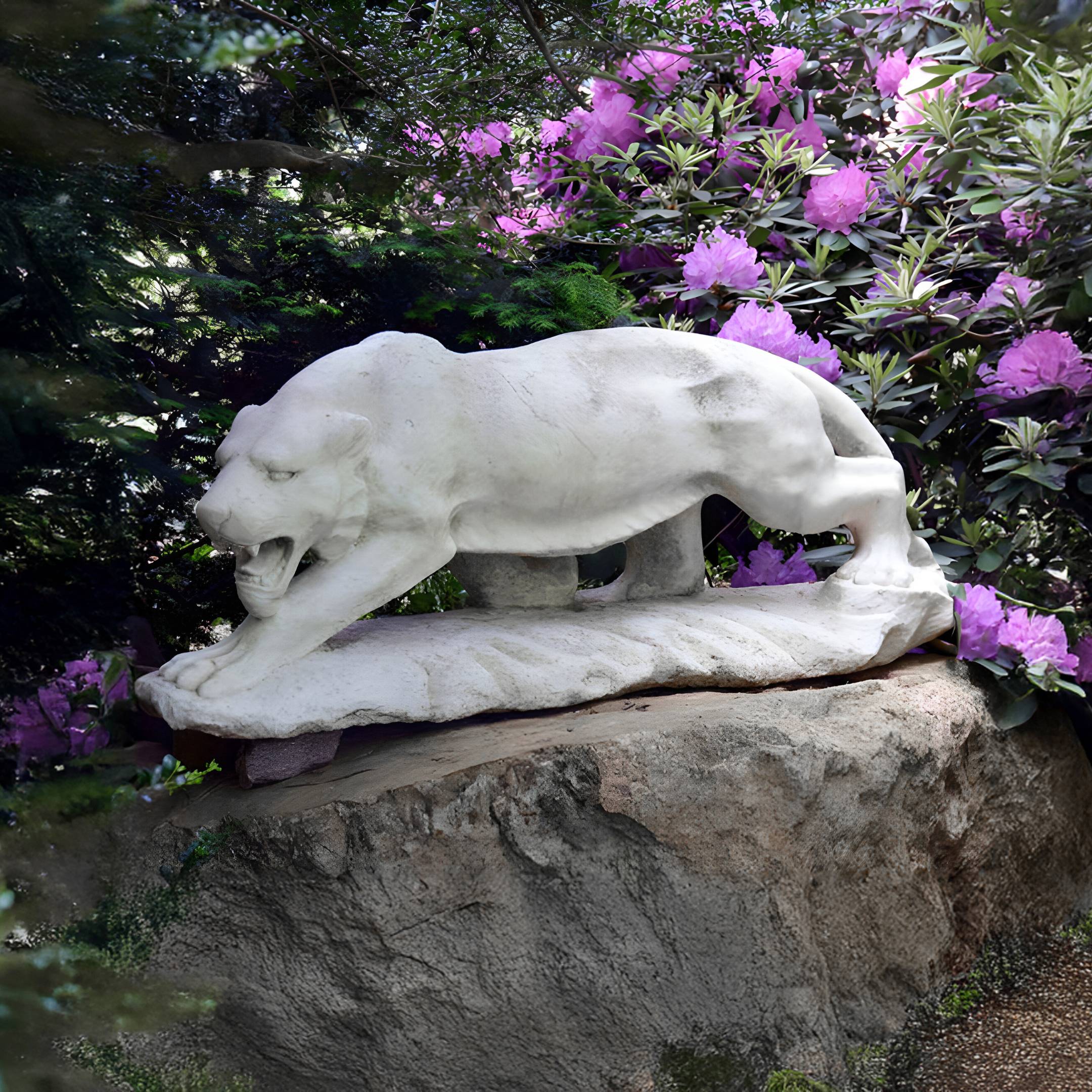 The Prowling Panther Marble Outdoor Sculpture - 65cm by Giant Sculptures stands majestically on a large rock, with its mouth open, surrounded by lush greenery and vibrant purple flowers, creating a natural and serene setting in your garden.